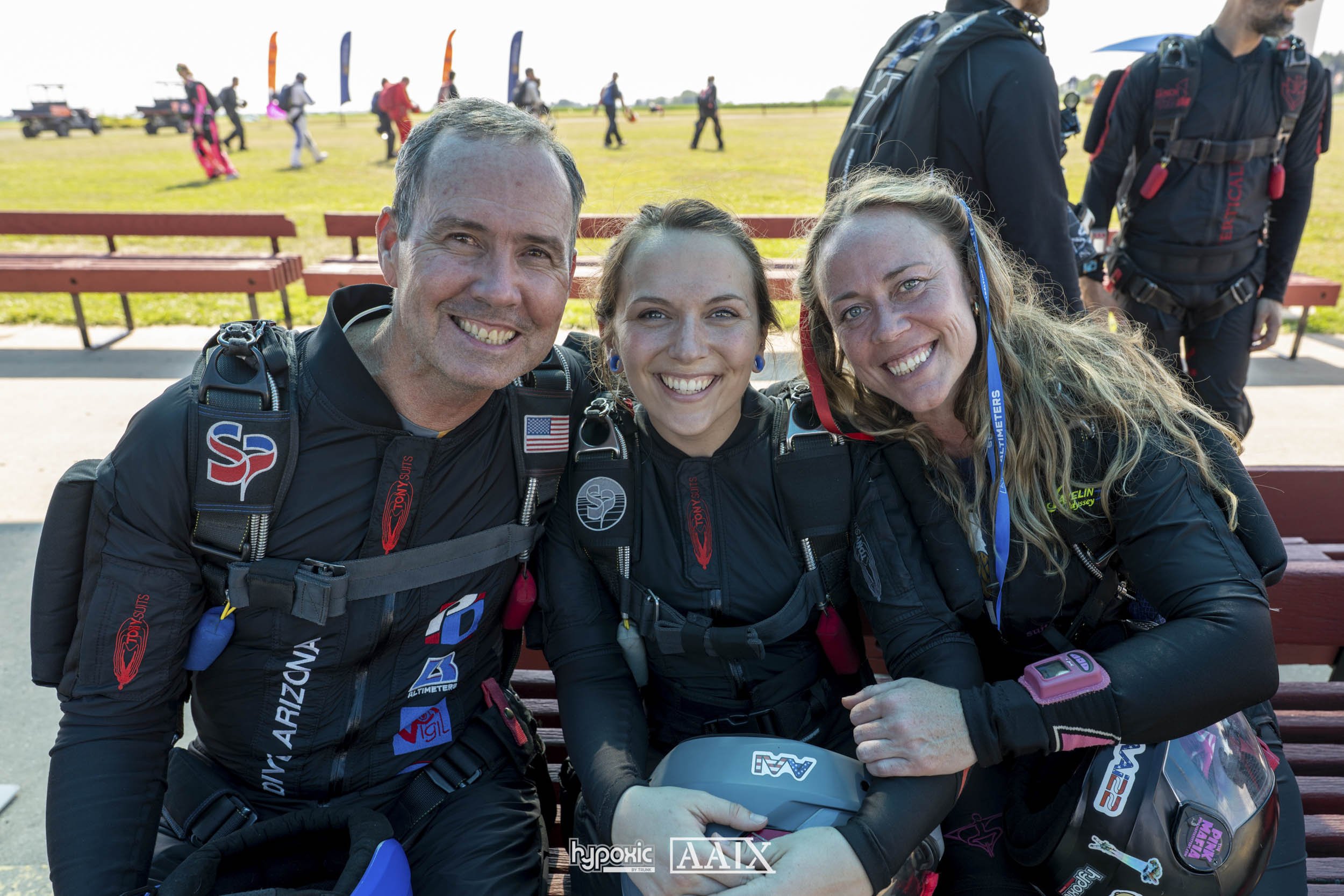 Three smiling skydivers sitting on a bench, wearing jumpsuits and gear, with a group of skydivers and open fields in the background.