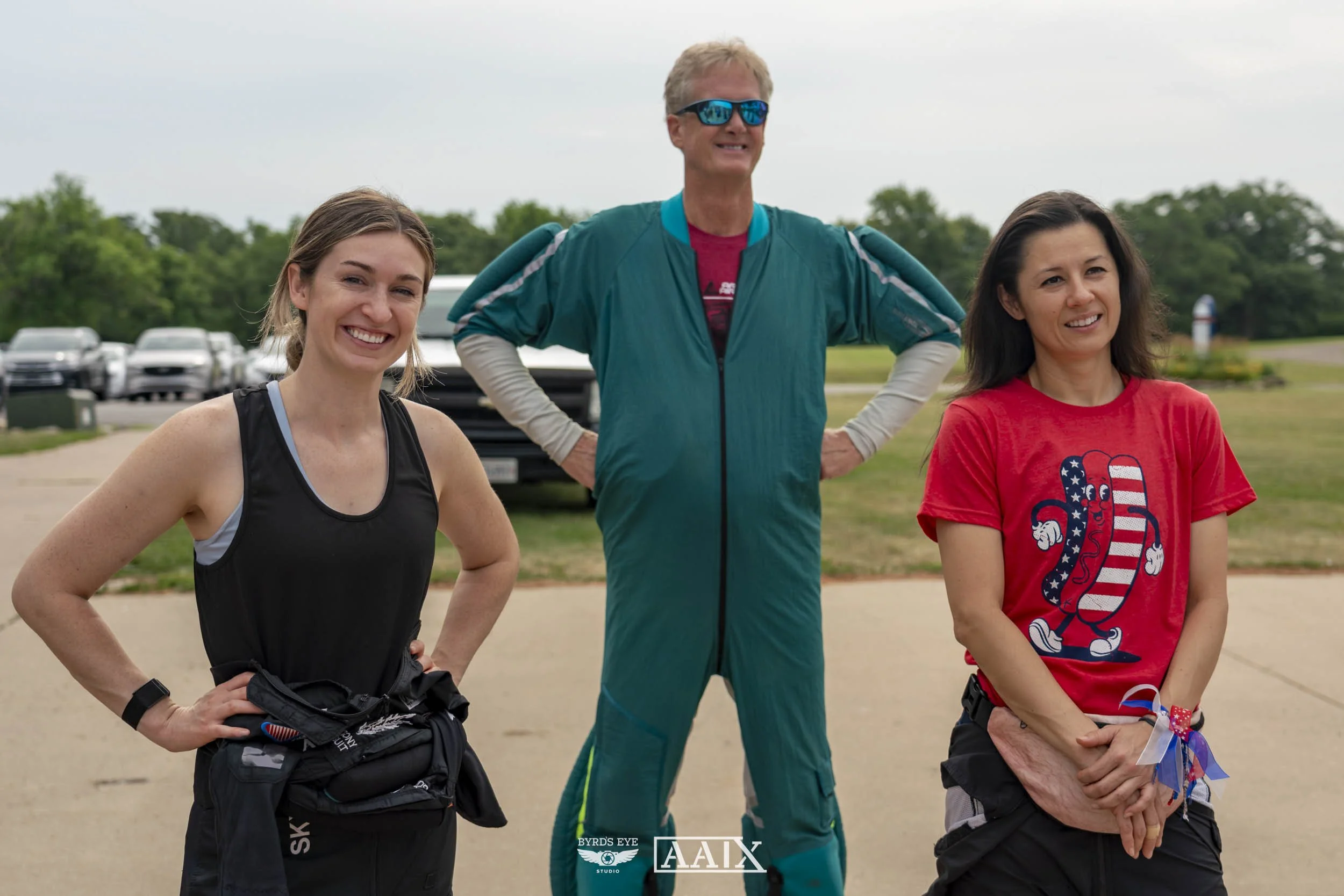 Three people, two women and one man, standing outdoors on a cloudy day. The woman on the left is wearing a black tank top and black pants, smiling with hands on hips. The man in the middle is wearing sunglasses, a teal jumpsuit with a red shirt under