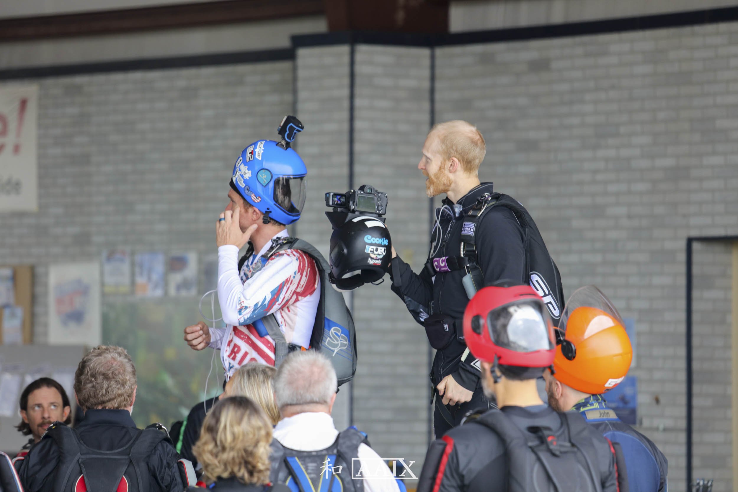 Motorsports competitor in racing gear with helmet, talking to a crew member holding a camera, surrounded by other team members and spectators, in a garage or pit area.