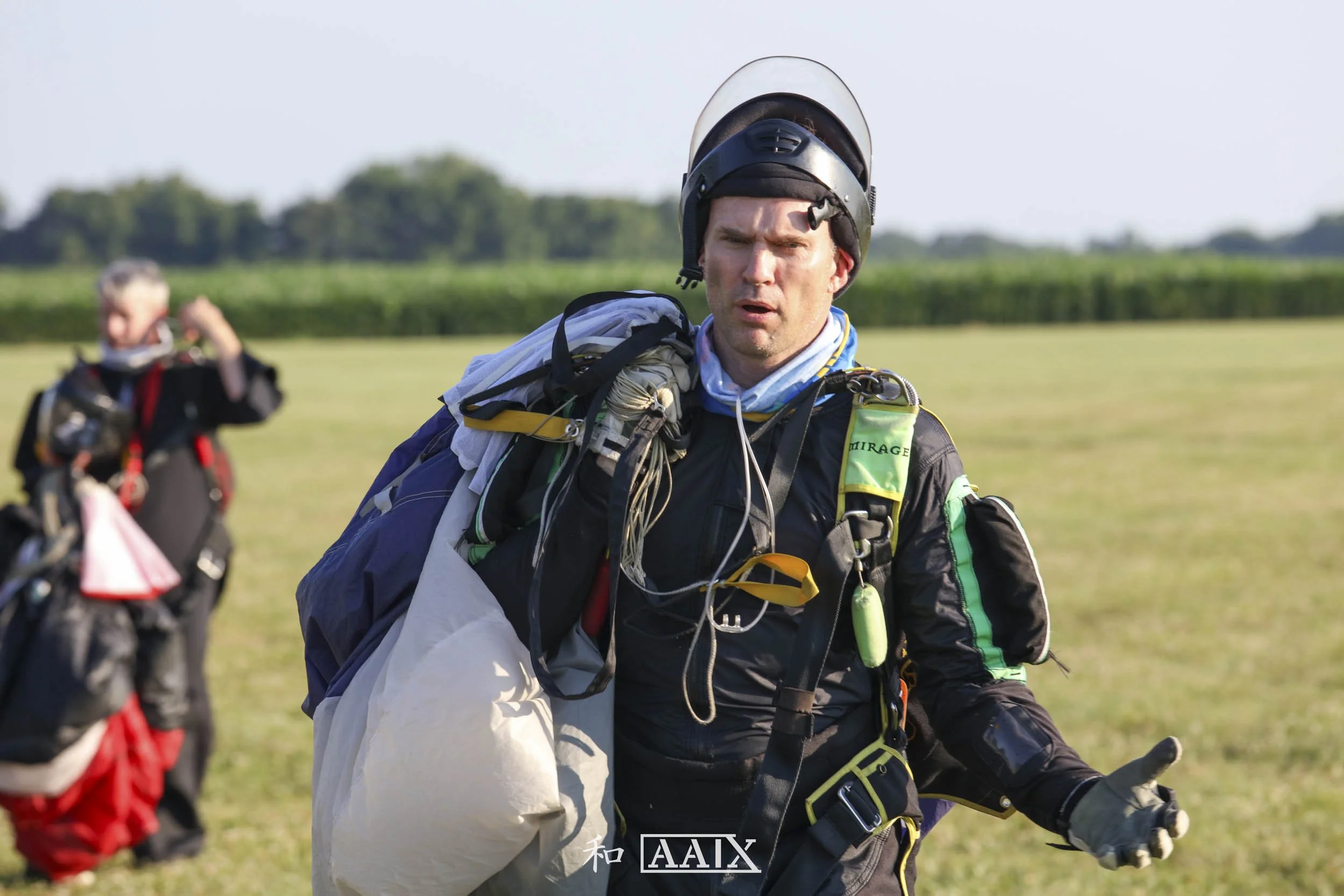Skydiver with helmet and jumpsuit, holding a parachute, on an open field, with another skydiver in the background.