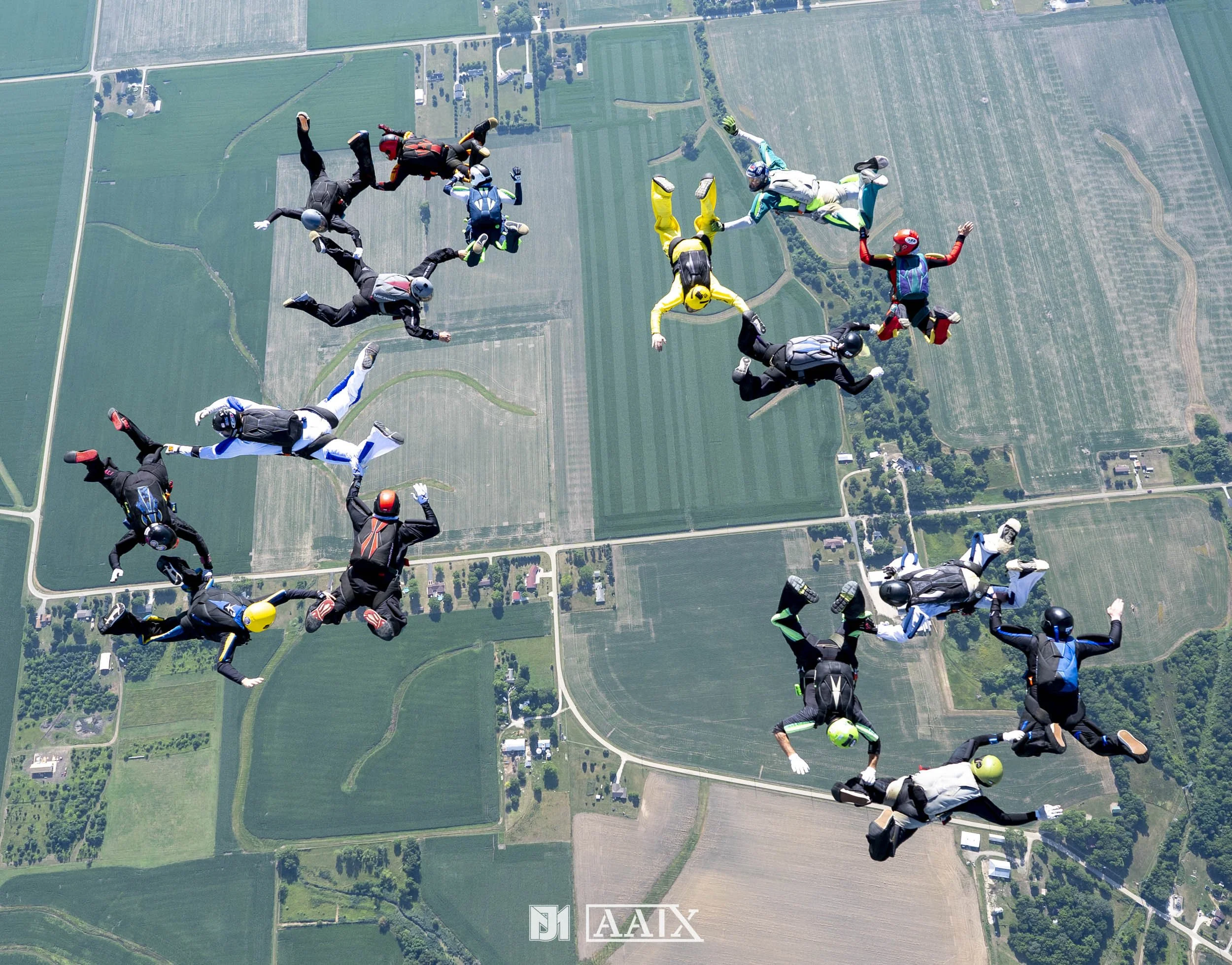 A group of skydivers in freefall over a patchwork of farmland and rural roads, each wearing jumpsuits and helmets, with the landscape below showing fields and small buildings.