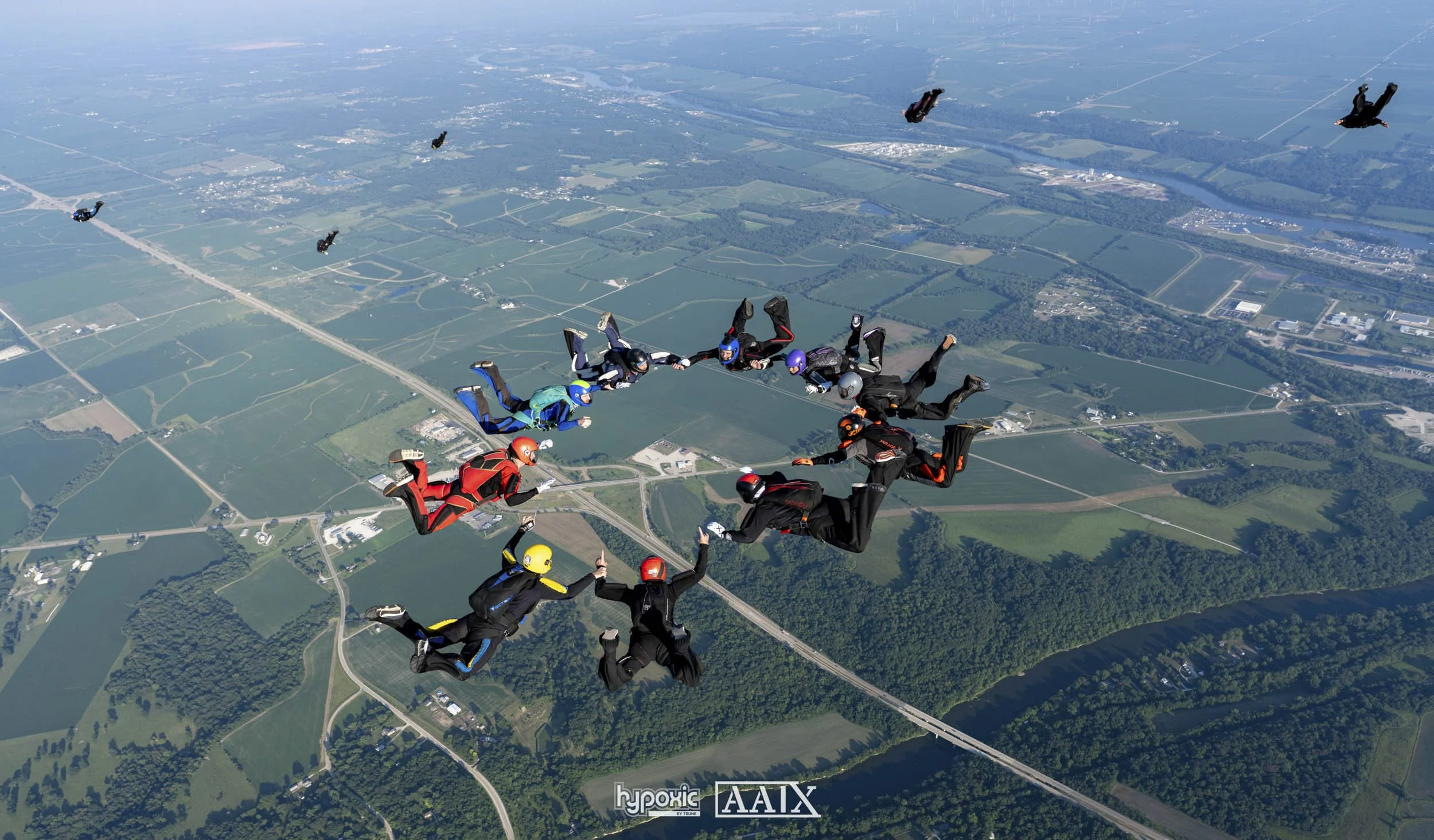 Group of skydivers in free fall over a rural landscape with fields, roads, and a river, wearing colorful jumpsuits and helmets.