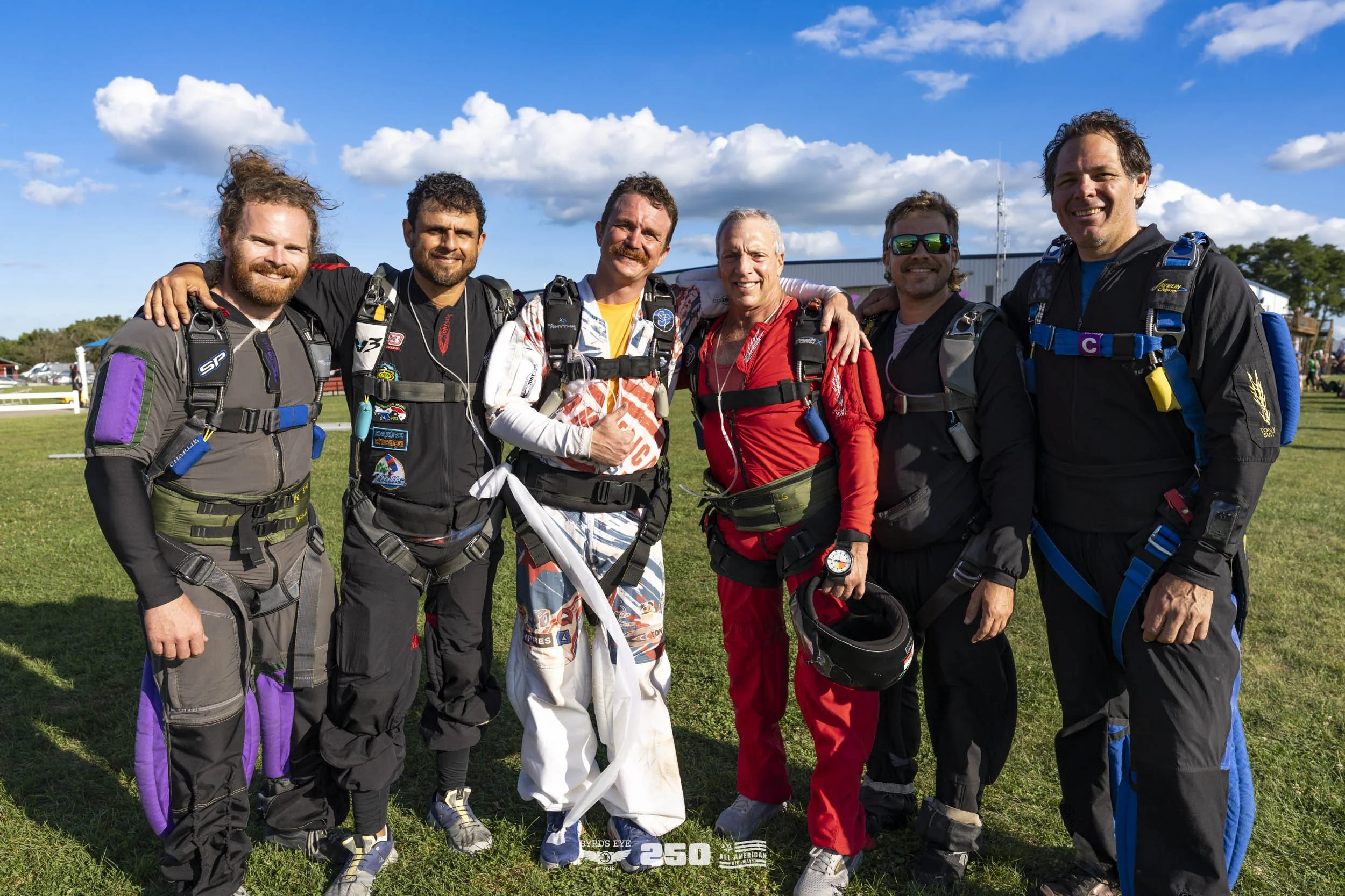 Seven skydivers standing together on grass, dressed in skydiving gear, smiling at the camera.