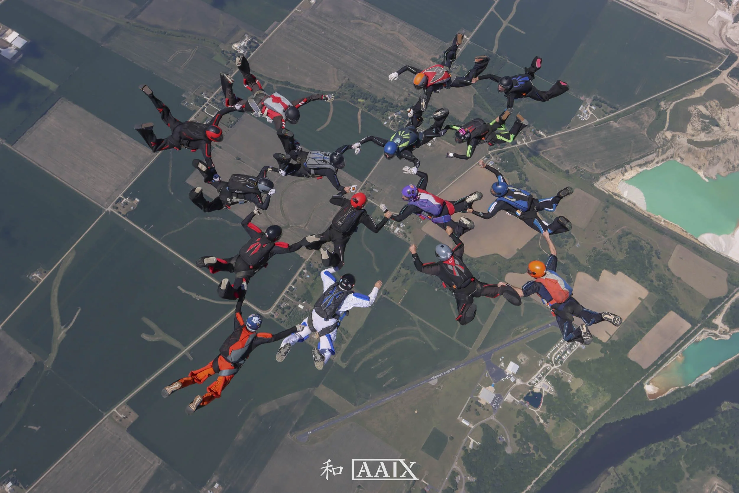 Group of skydivers in freefall formation, holding hands, with a landscape of fields, lakes, and roads below.
