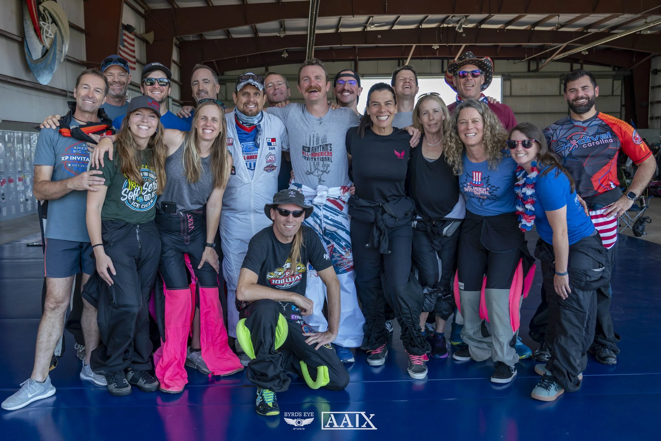Group of people smiling and posing indoors, some in athletic gear, in a large hangar-like space.