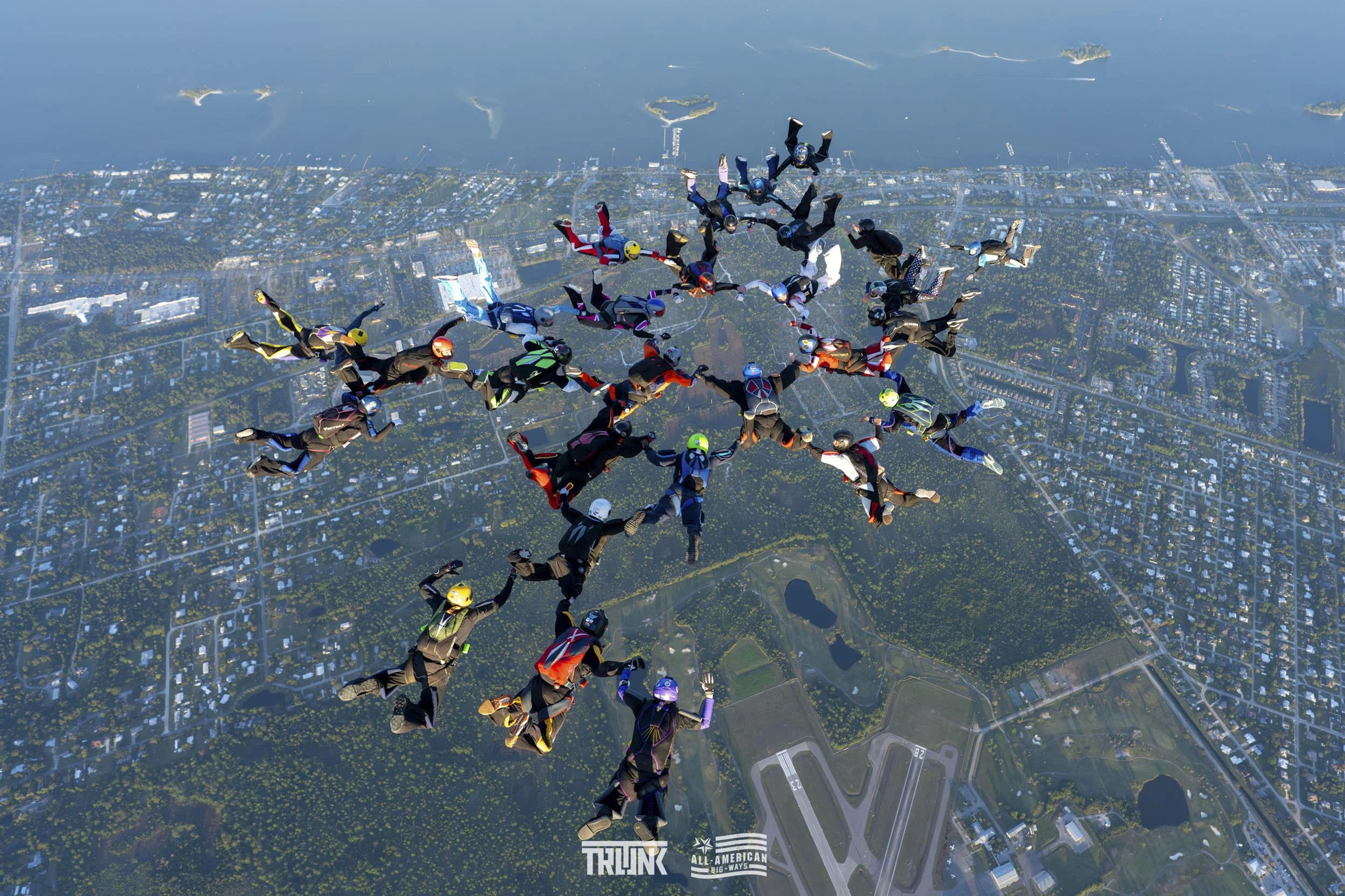 Group of skydivers holding hands forming a star shape during freefall over a city with lakes and parks.