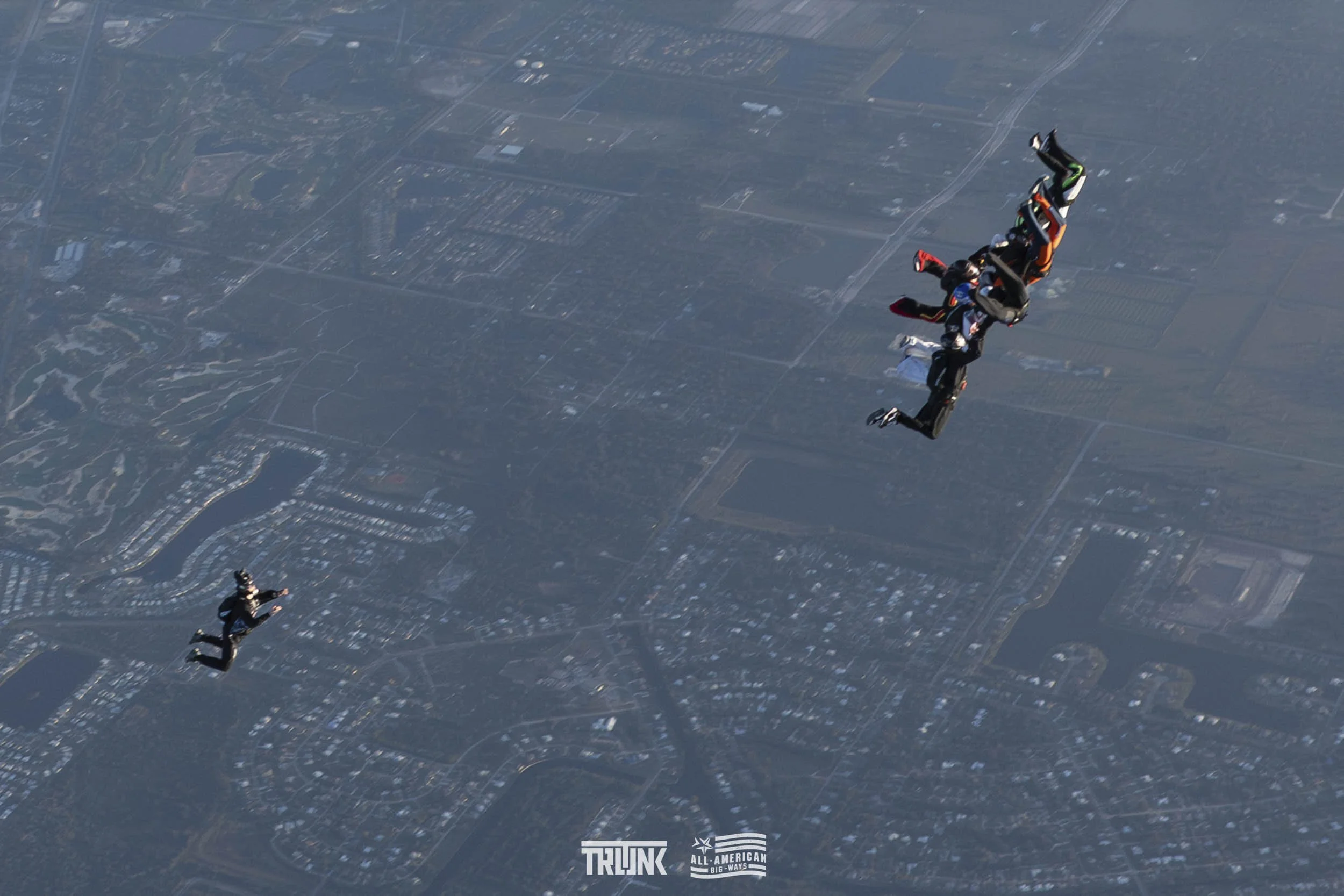 Five skydivers in freefall formation over a city landscape during daytime.