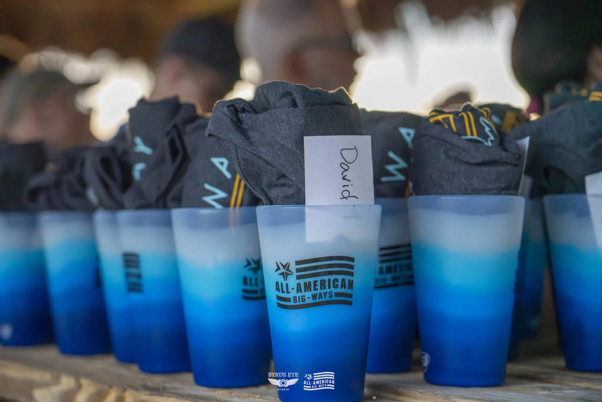 A row of blue and white gradient cups with the All-American Big-Ways logo, containing folded dark clothing items with a label reading 'David.'