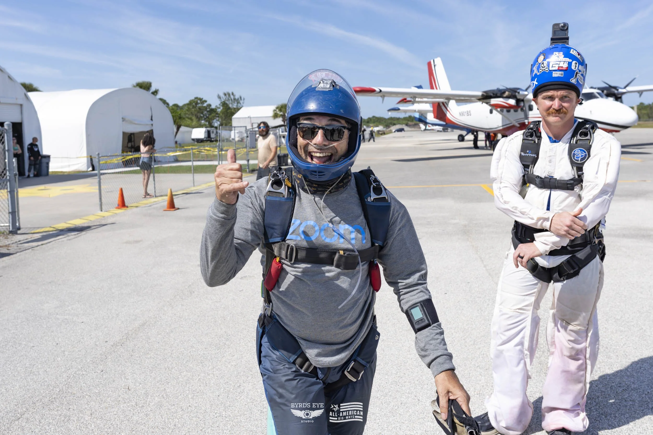 Two skydivers in jumpsuits and helmets standing on an airfield, with a plane in the background, one smiling and giving a thumbs-up.