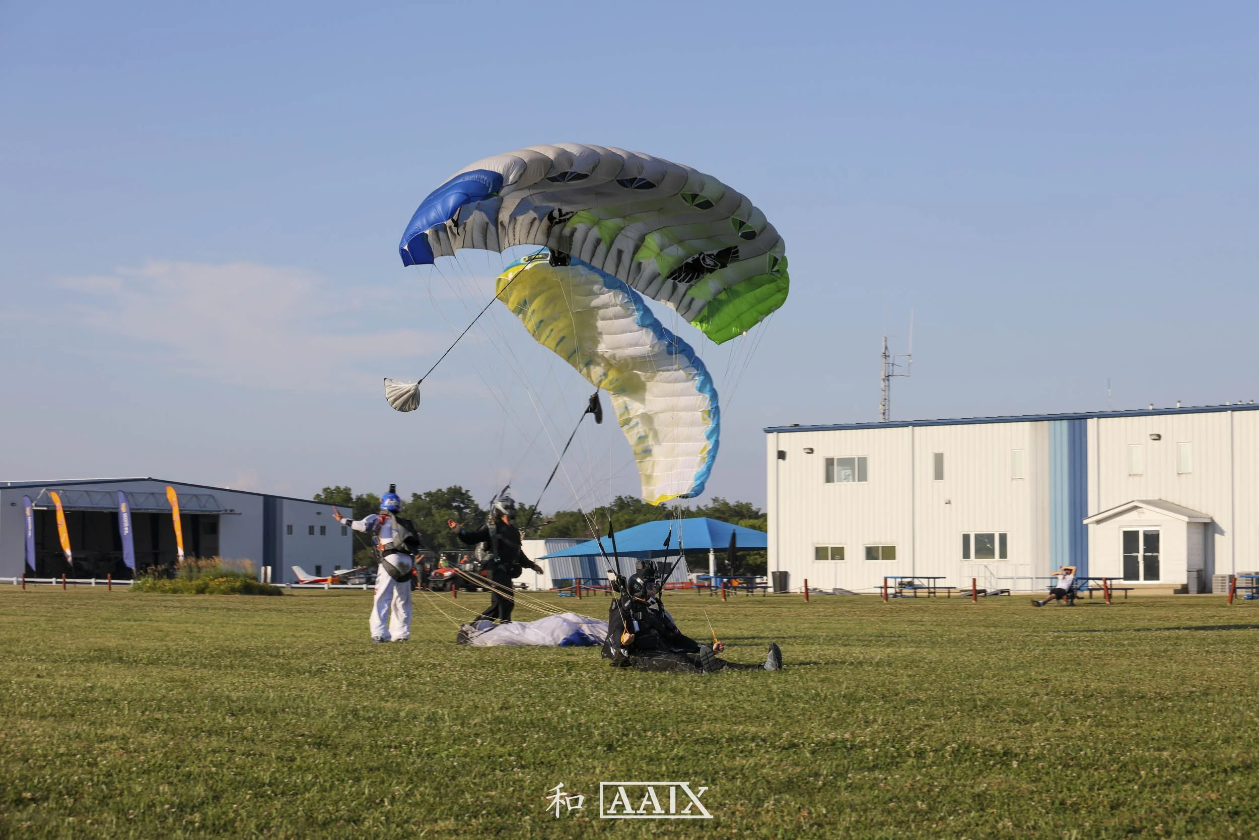Two paragliders with colorful parachutes preparing for takeoff on a grassy field, with a few spectators and buildings in the background under a clear sky.