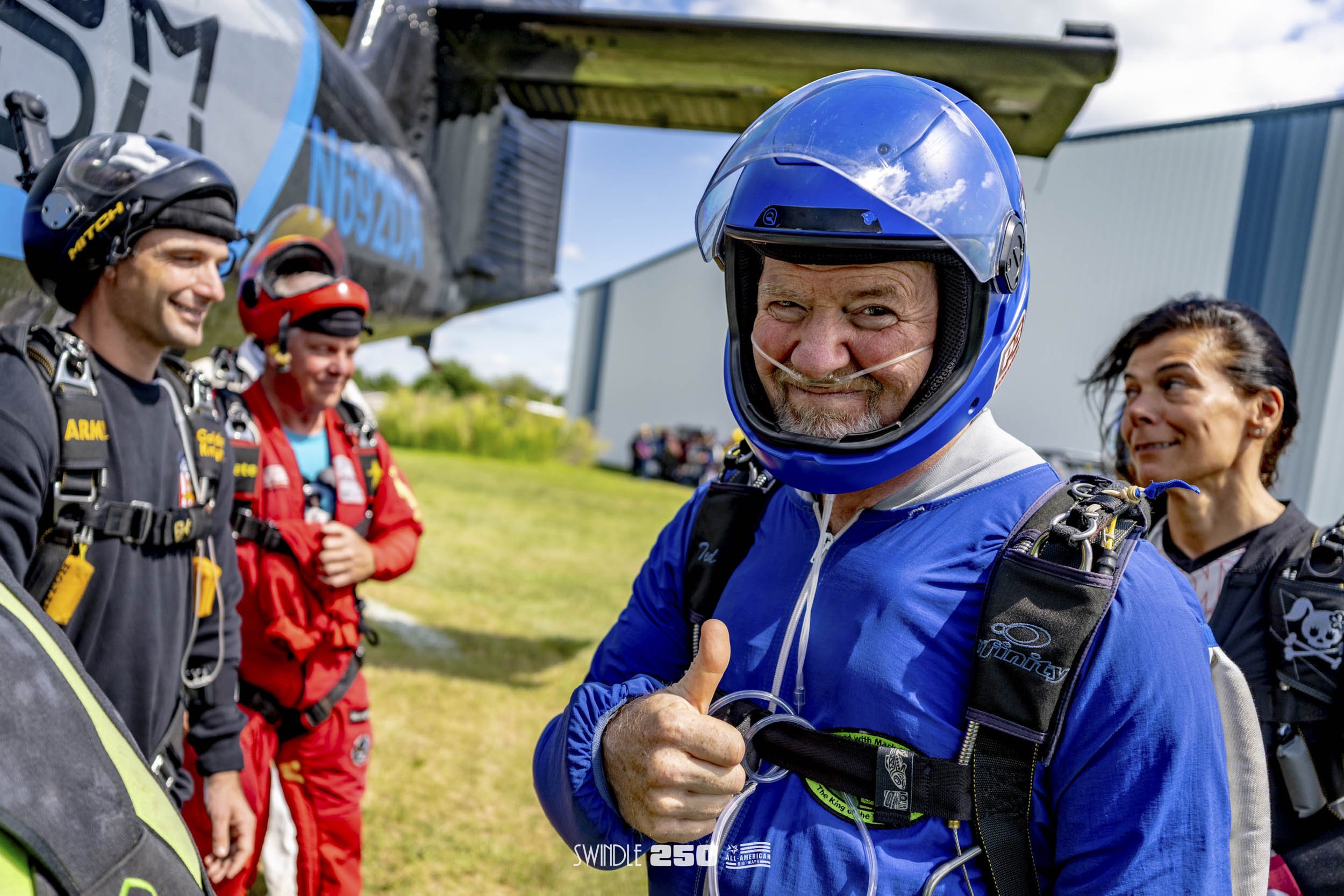 Skydivers preparing for a jump, with one smiling man wearing a blue jumpsuit and helmet giving a thumbs-up, and others in the background near an aircraft on a grassy field.