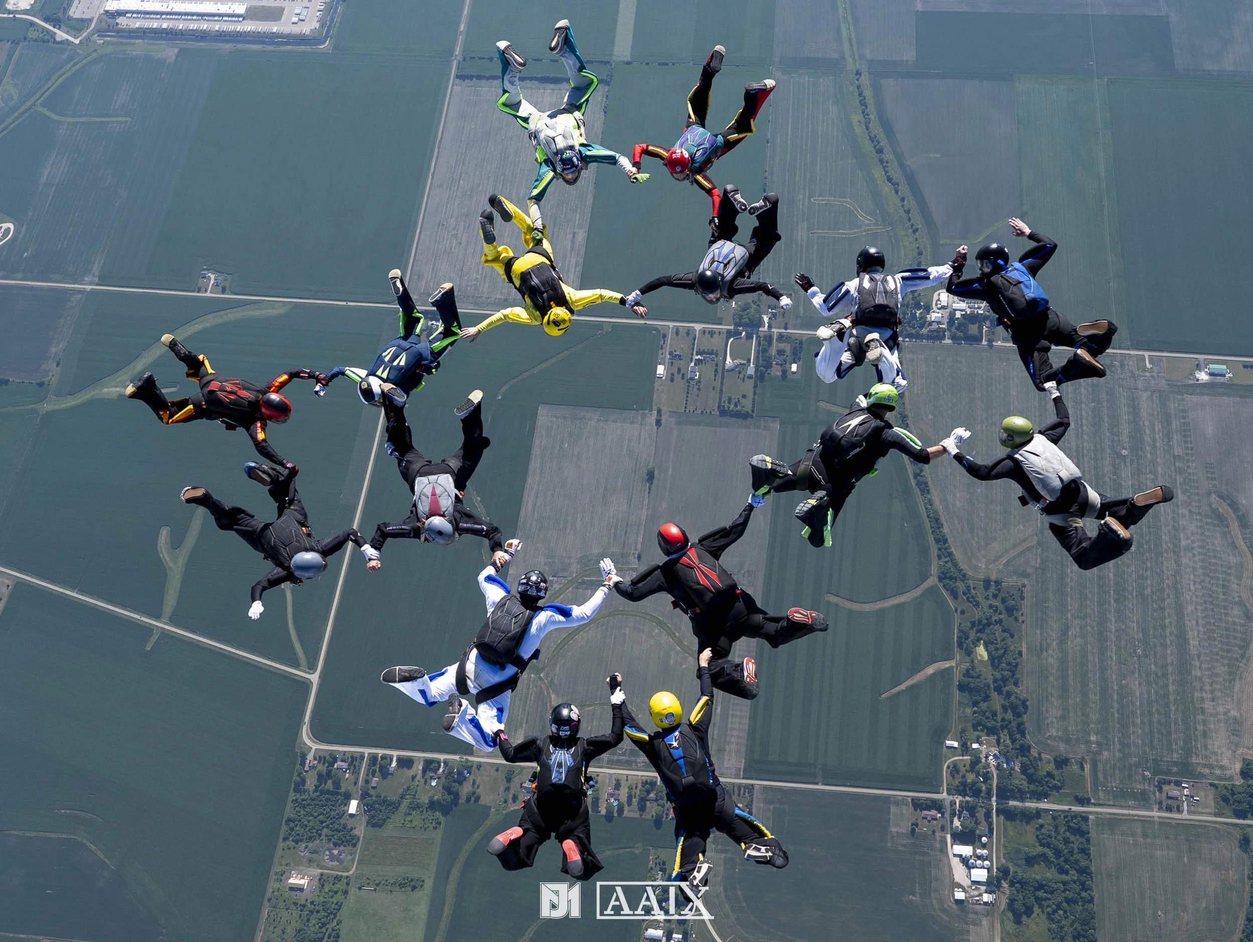 Group of skydivers forming a star shape in mid-air above farmland, holding hands and wearing colorful jumpsuits and helmets.
