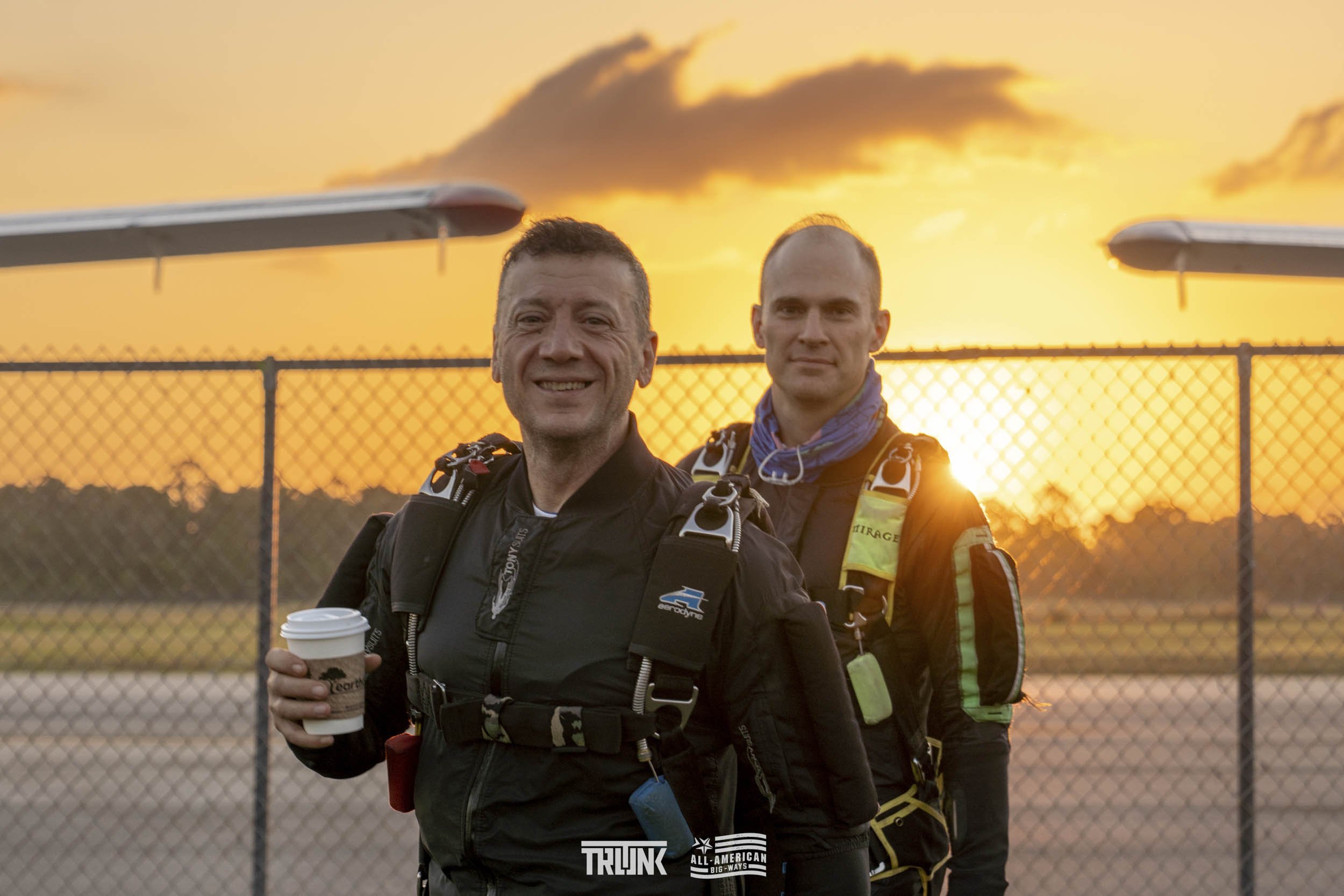 Two skydivers in gear smiling at sunset, with a chain-link fence and clouds in the background.