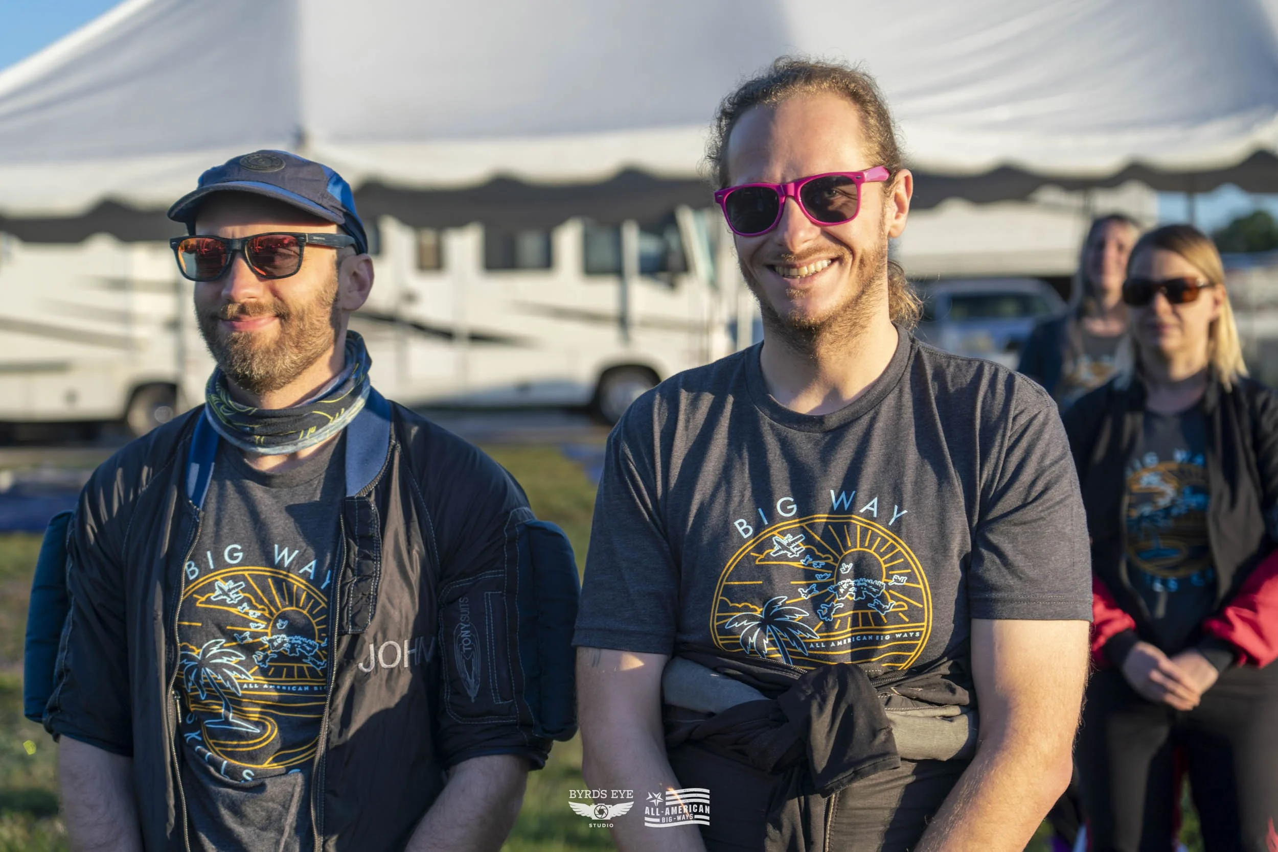 Group of four people outdoors wearing matching "Big Way" t-shirts, some with sunglasses. Two men in the front, smiling, with a white tent and RV in the background.