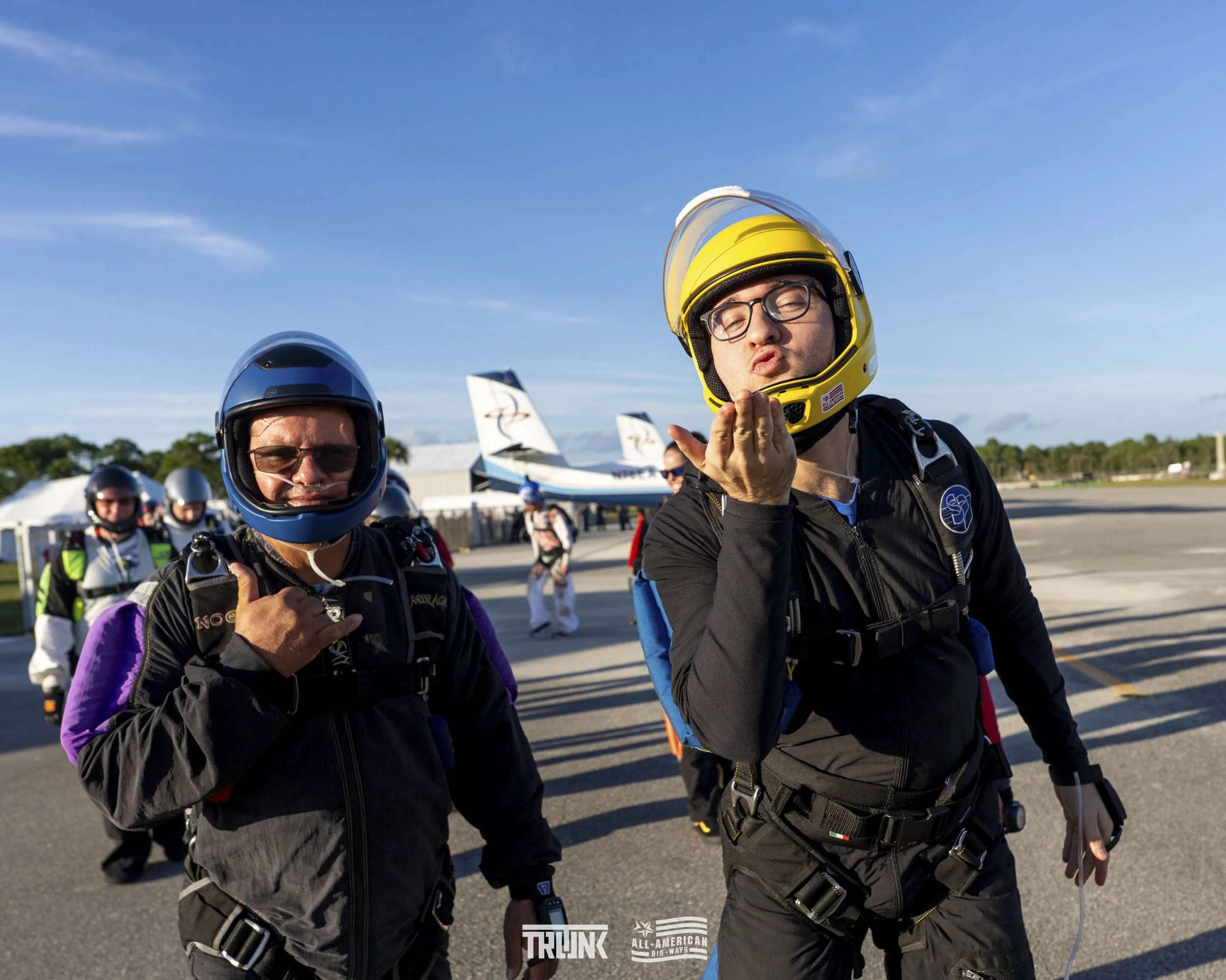 Two skydivers in jumpsuits and helmets on an airstrip with planes in the background. The person on the right is blowing a kiss, and the person on the left is holding his harness.