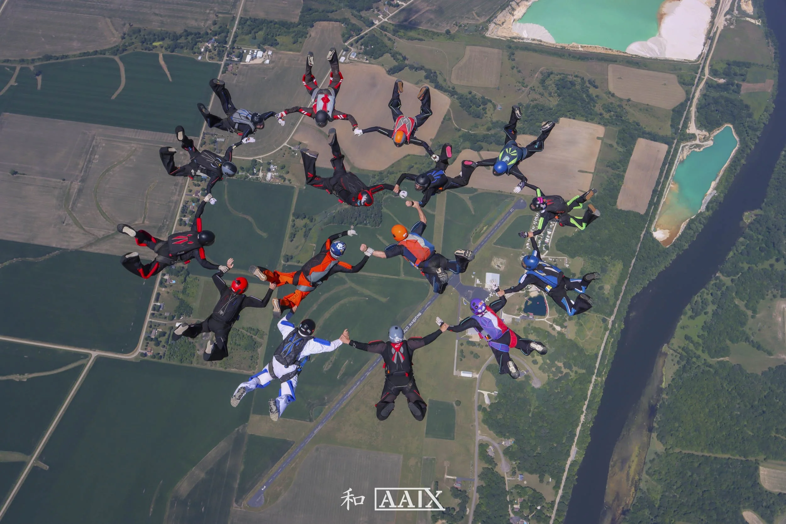A group of skydivers in colorful suits and helmets forming a circle while mid-air over a patchwork landscape of fields, roads, a river, and ponds.