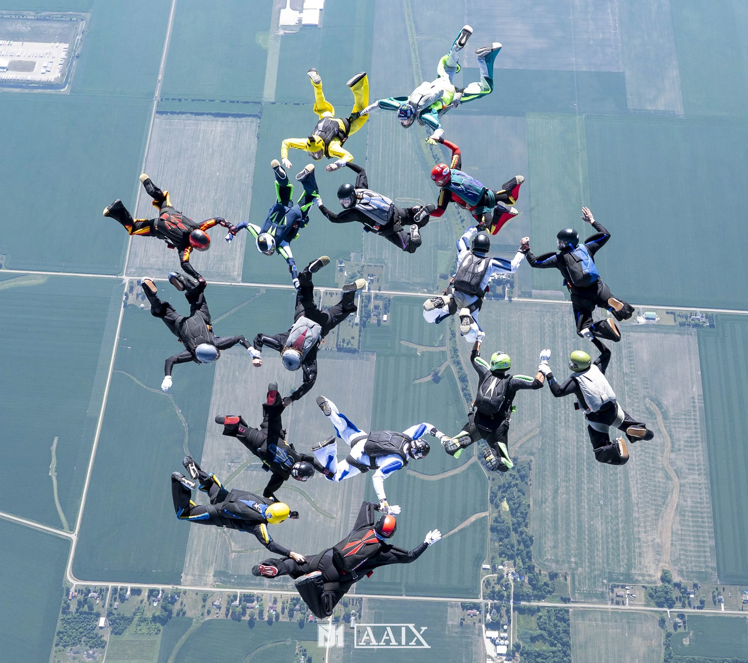 Group of skydivers forming a human chain while free-falling over a farmland landscape.