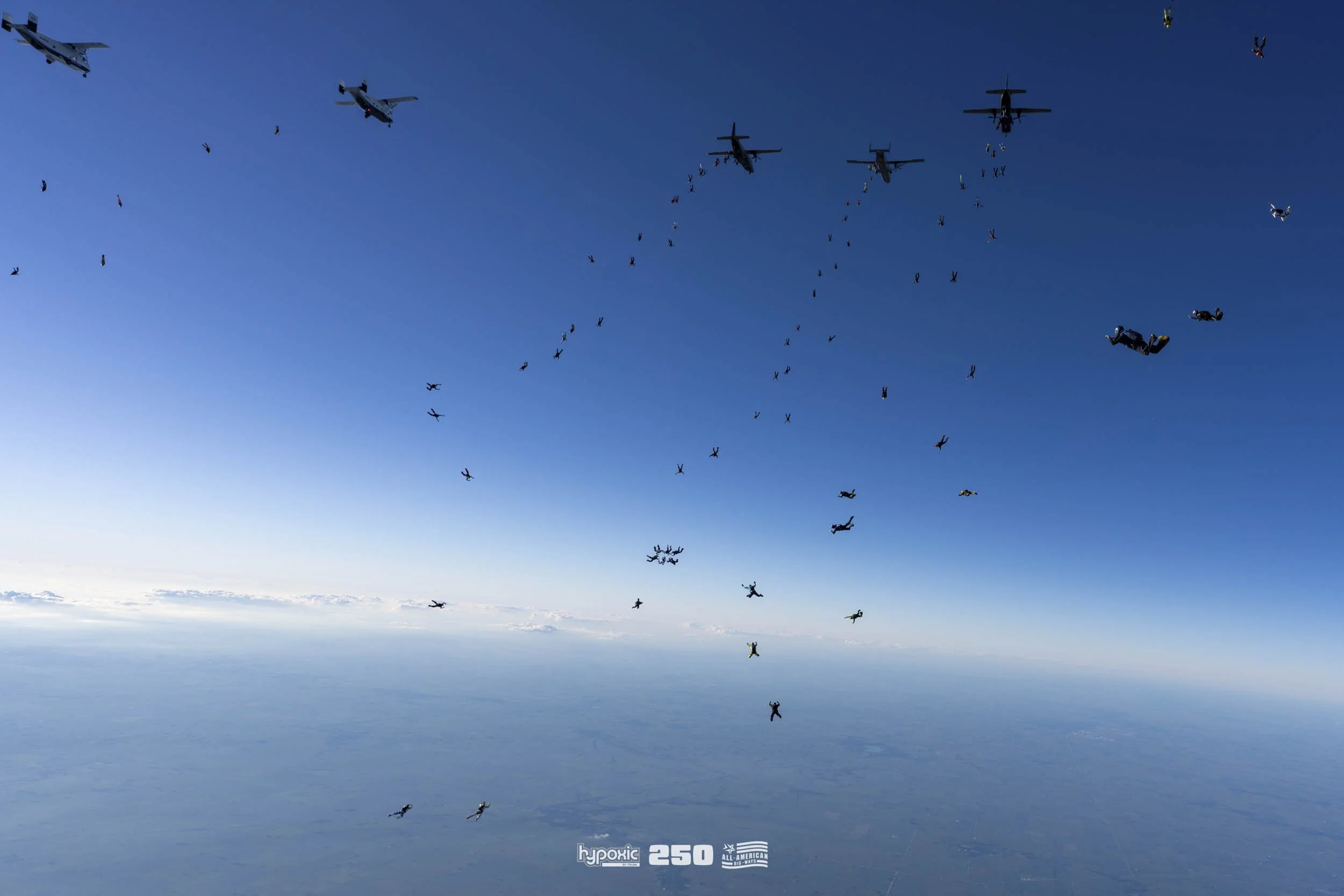 Skydivers in free fall formation above the clouds with four military aircraft flying overhead against a clear blue sky.