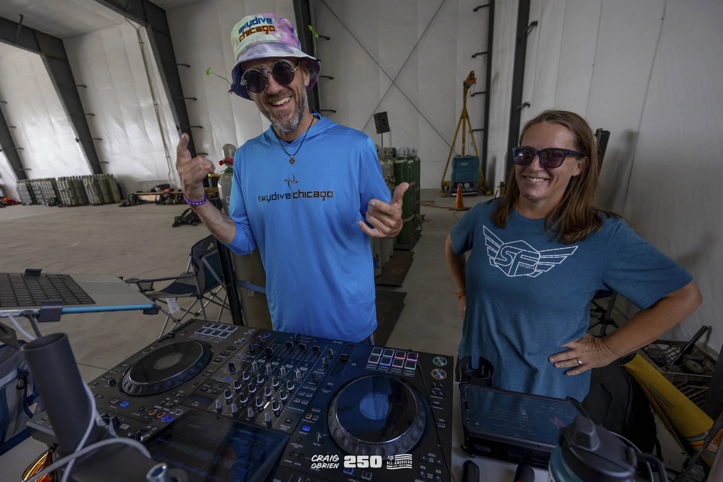 Two people smiling and standing behind DJ equipment inside a tent. The man on the left wears a colorful bucket hat, sunglasses, and a blue long-sleeve shirt that says 'eskydive chicago'. The woman on the right wears sunglasses and a blue T-shirt with