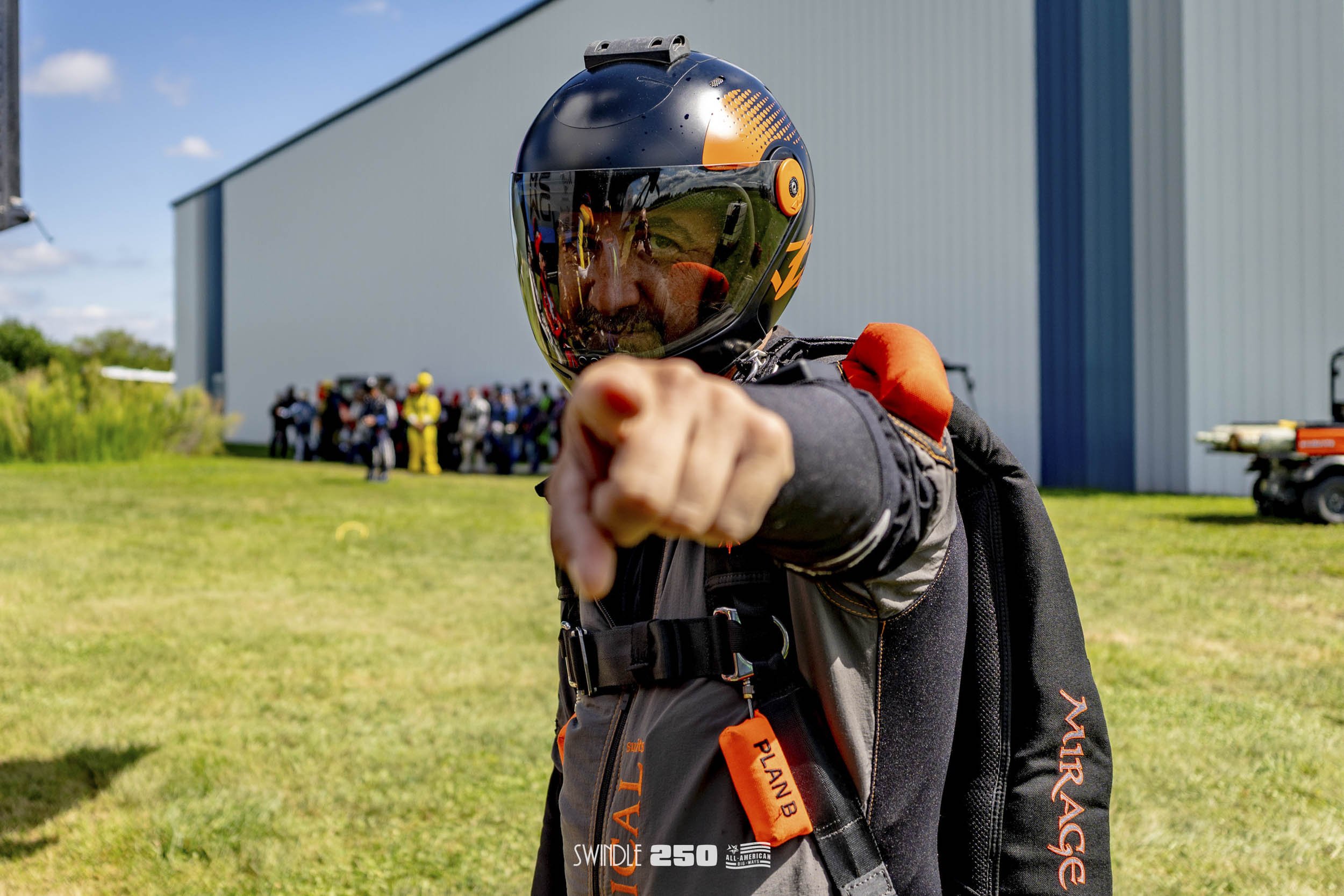A skydiver wearing a helmet and jumpsuit, pointing towards the camera with a group of skydivers and a building in the background.