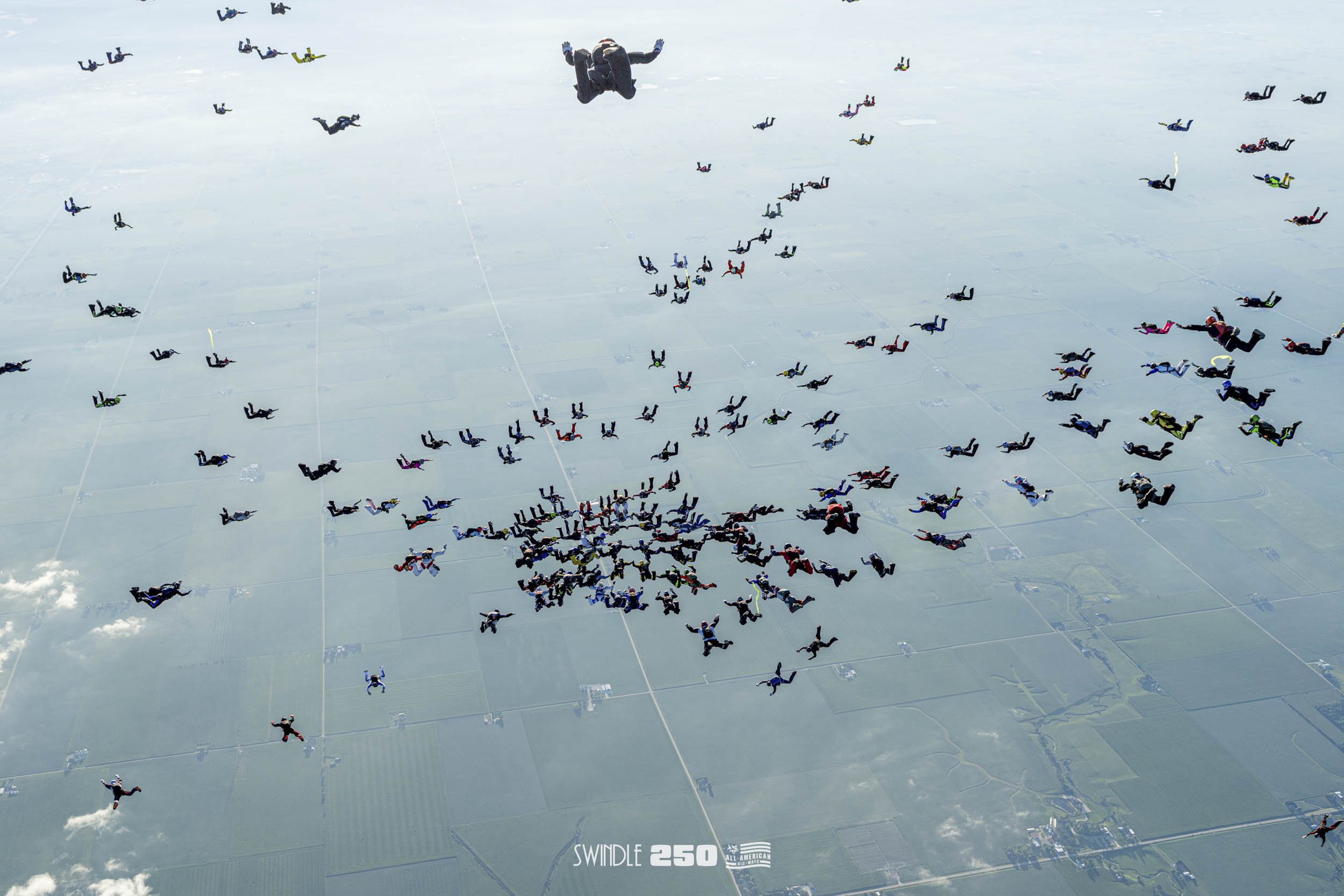 A large group of skydivers in colorful jumpsuits and parachutes mid-air during a coordinated jump over a landscape with fields, and the word 'SWINDLE 250' with the American flag logo at the bottom.