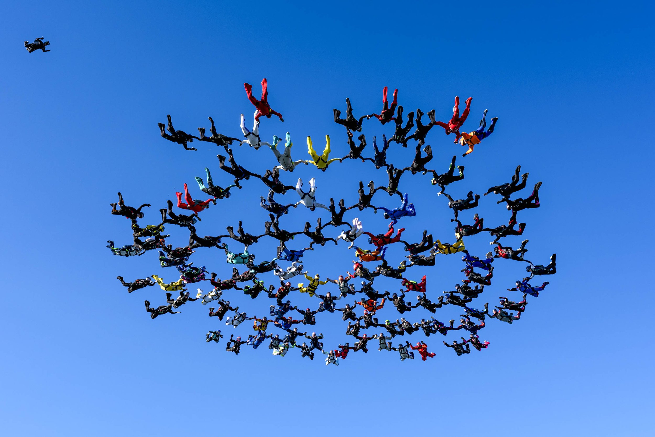 A large group of skydivers in colorful jumpsuits forming a cloud shape in the blue sky, with one skydiver in black in the top left corner.