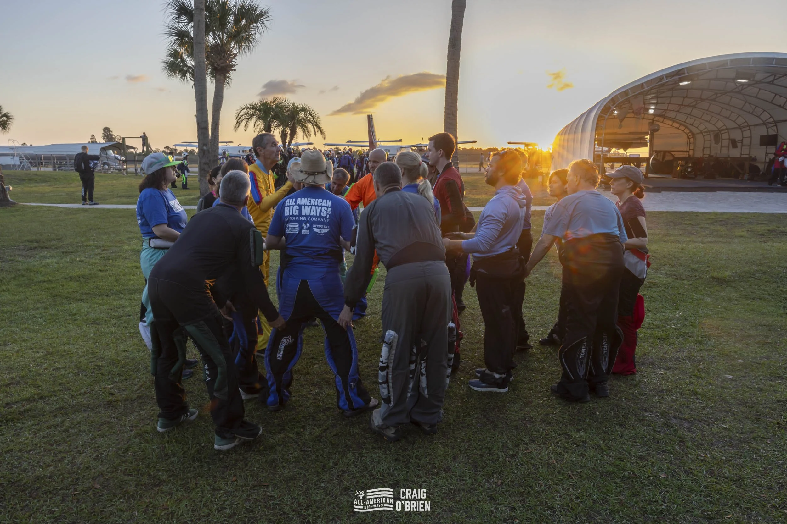 Group of people holding hands in a circle outdoors during sunset at an airfield.