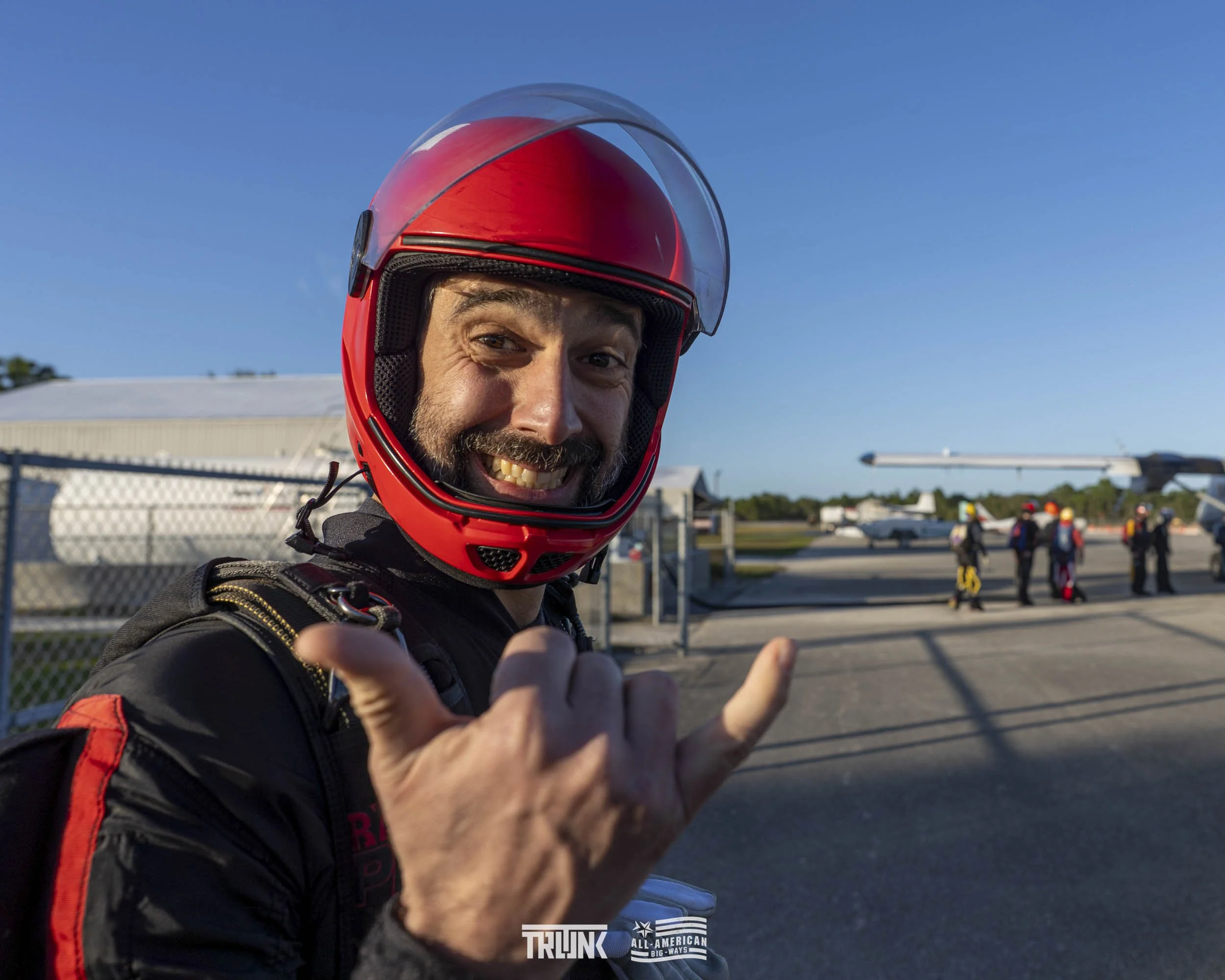 A man smiling and making a shaka sign while wearing a red motorcycle helmet and black motorcycle gear at an airport tarmac, with a group of people and aircraft in the background.