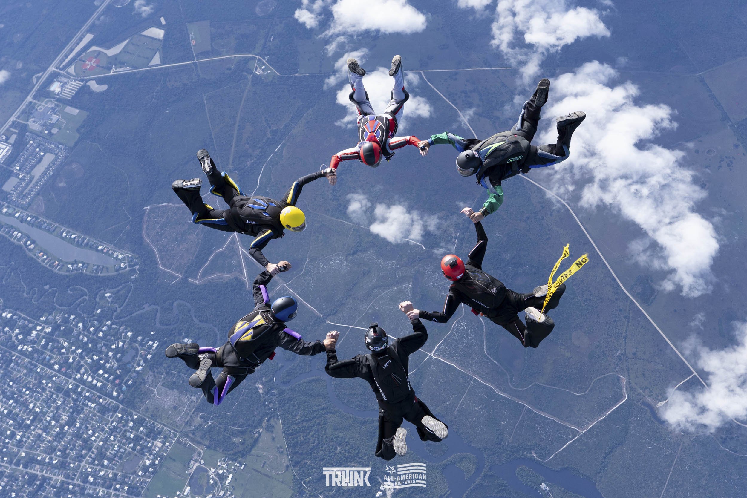 Group of skydivers holding hands during free fall over a landscape with clouds, forests, roads, and buildings.