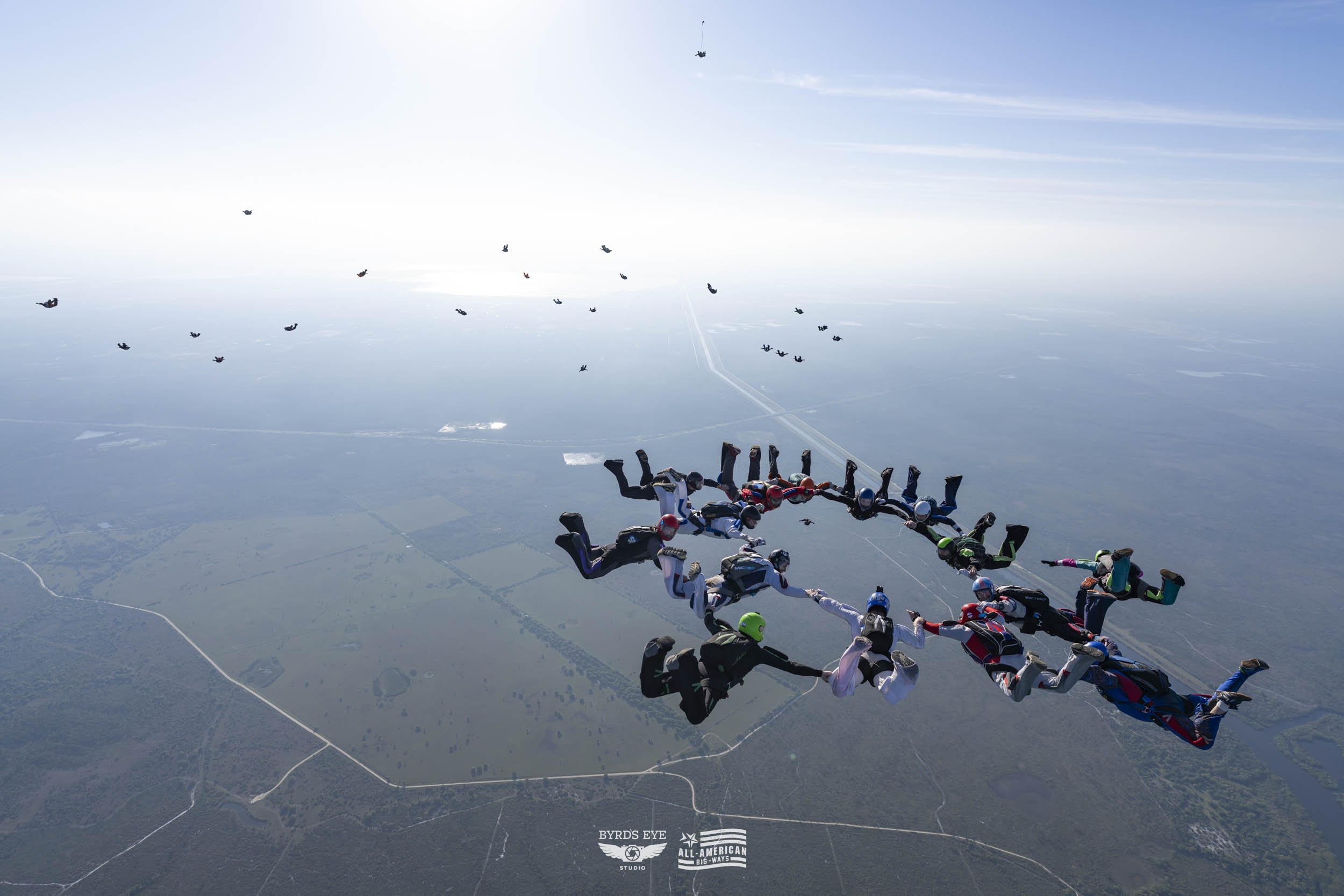 Multiple skydivers forming a circle while free-falling in the sky over a landscape.