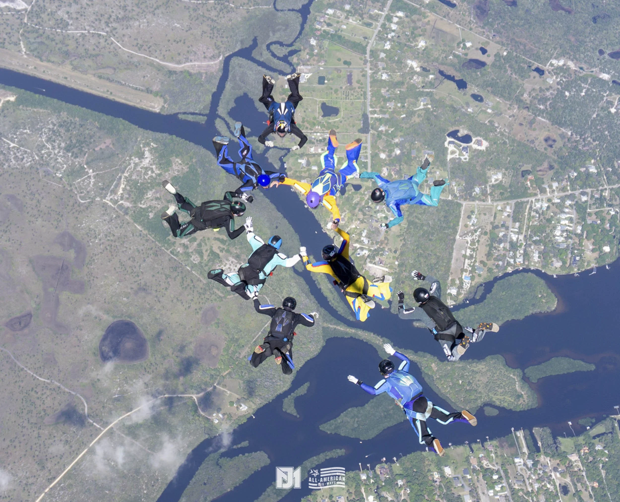 Multiple skydivers in colorful jumpsuits freefall over a landscape with lakes, rivers, and a small town.