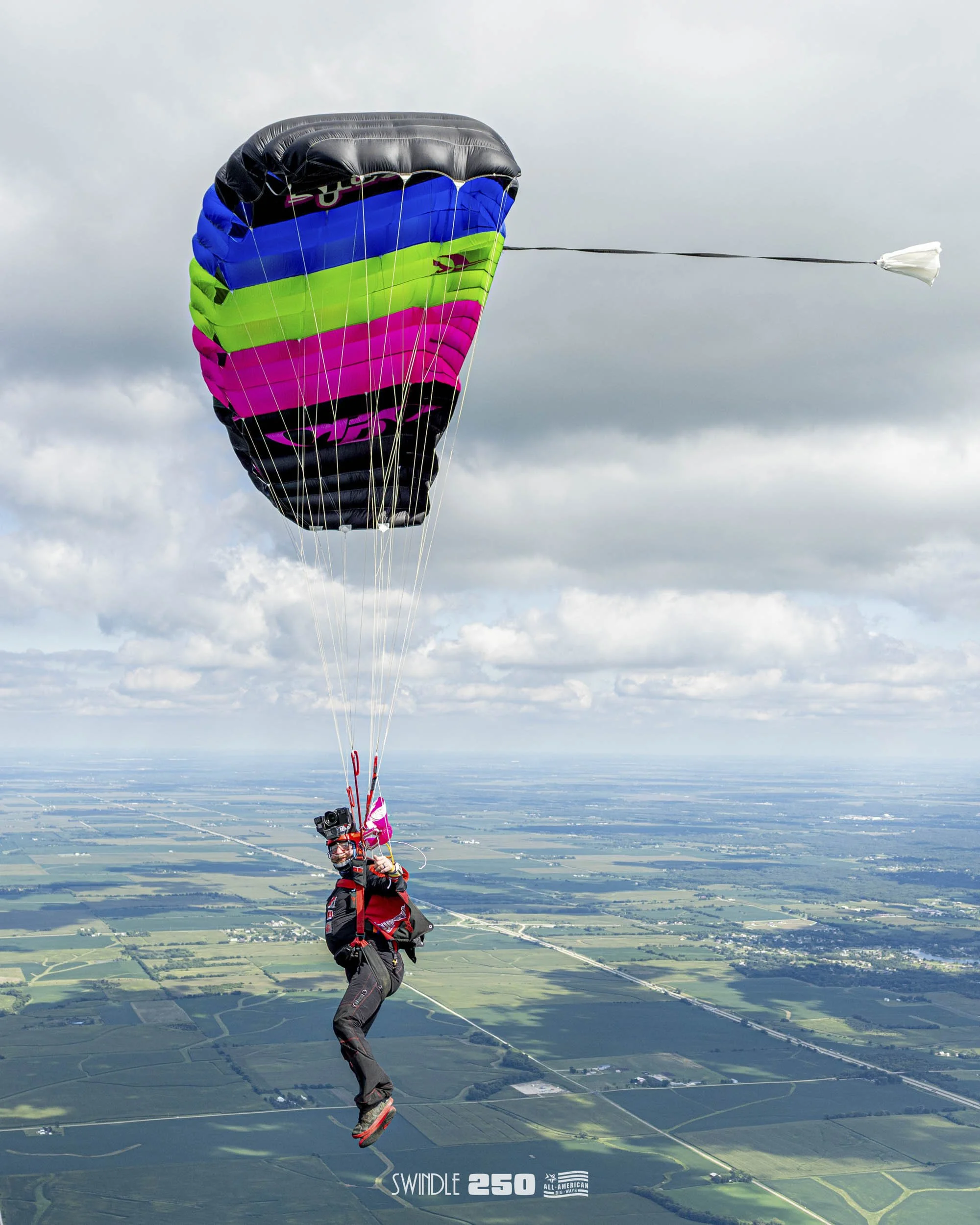 Paraglider flying high above a landscape with fields, wearing a red and black suit, and a colorful parachute with blue, green, pink, and black sections.