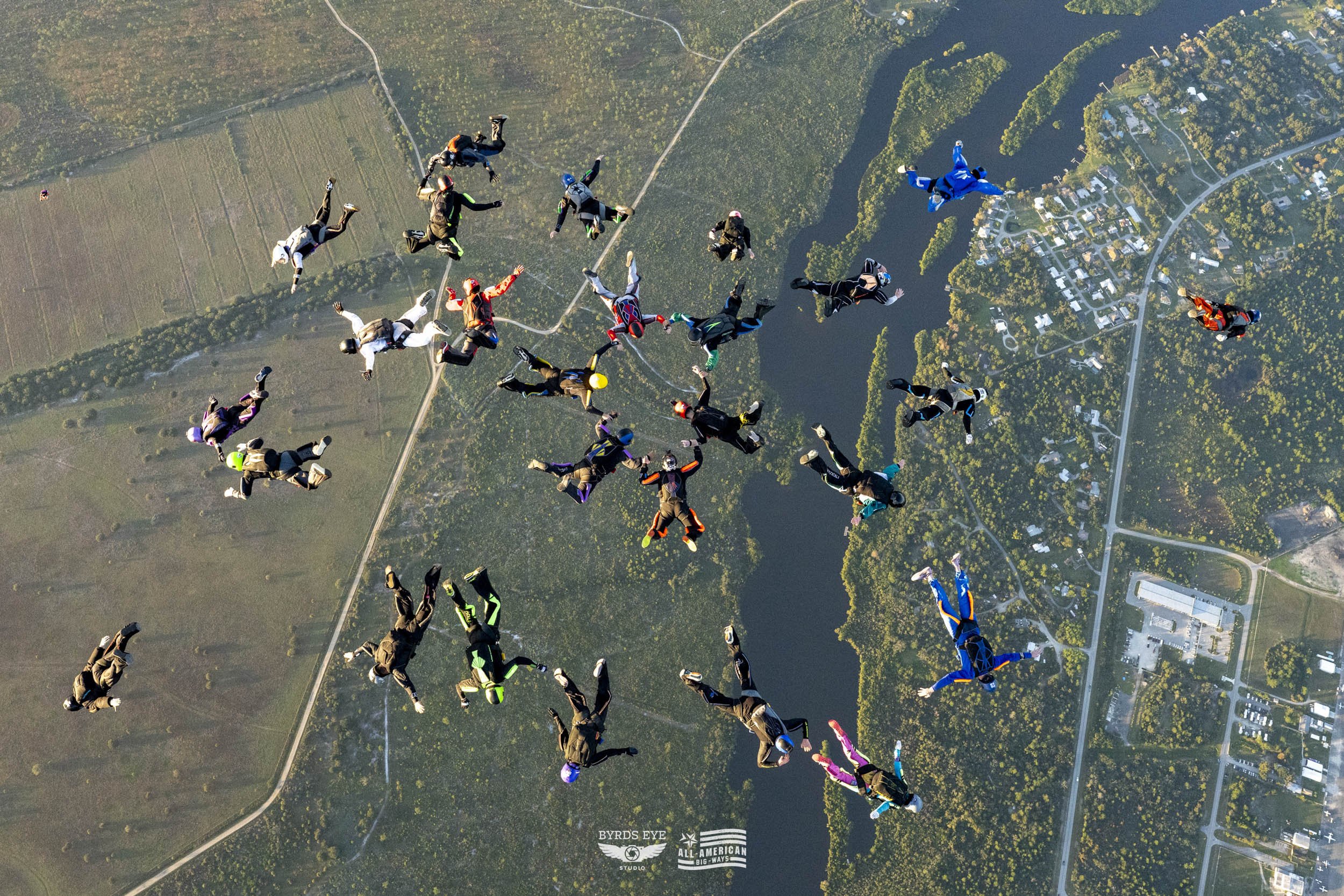 Group of skydivers in freefall formation above a landscape with water, trees, roads, and buildings.