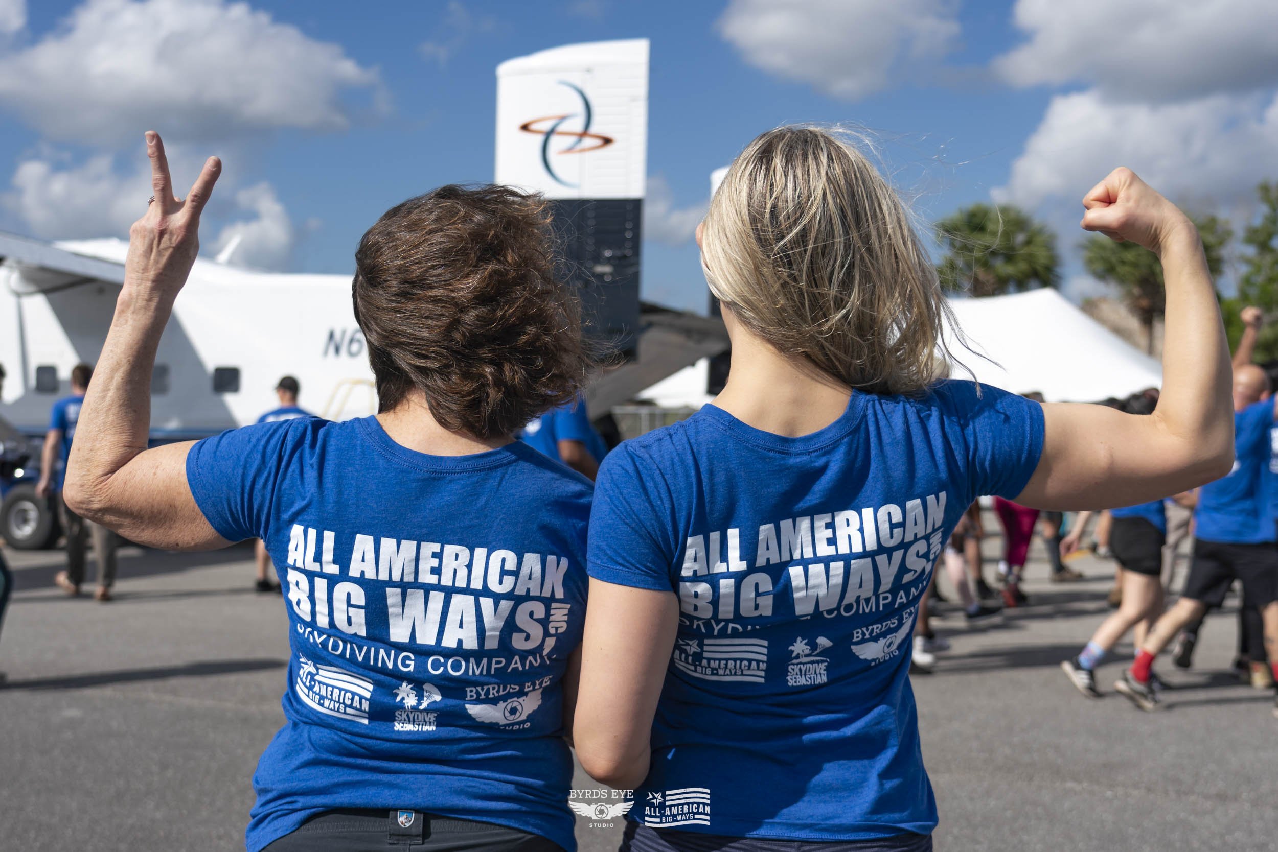 Two women with gray hair and wearing matching blue shirts displaying 'All American Big Ways Skydiving Company' print, flexing their arms and participating in an outdoor event with other people and a small aircraft in the background.
