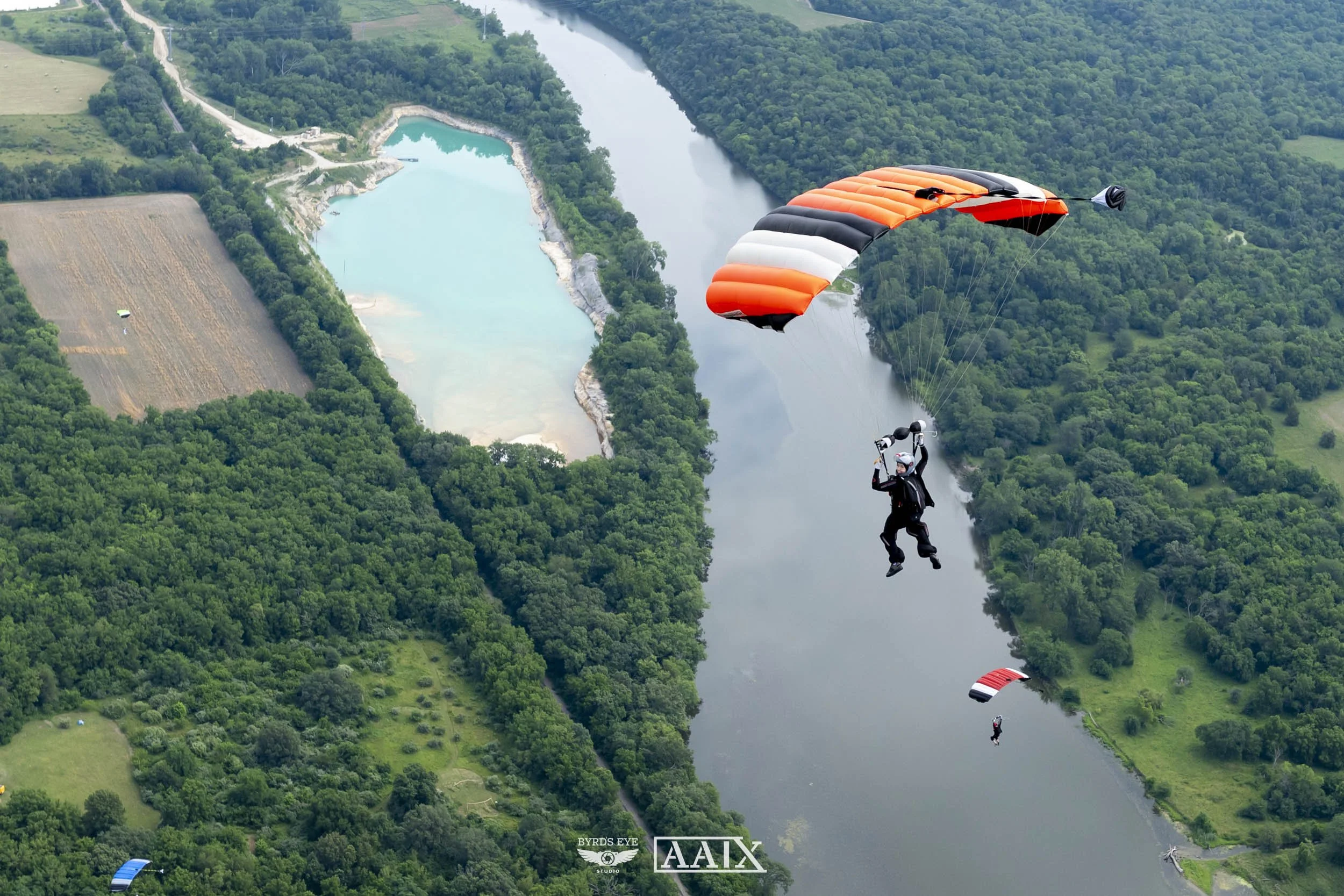 A person is skydiving over a river with a forested landscape and open fields surrounding it.