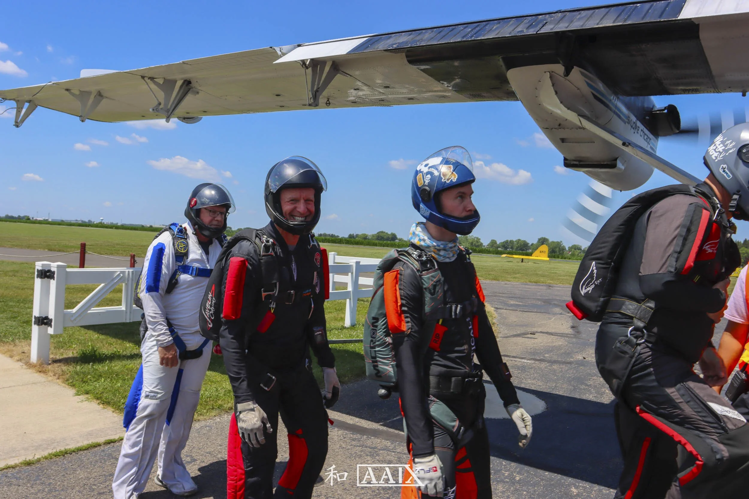 Four skydivers standing in front of an airplane, preparing for a jump on a sunny day at an airfield.