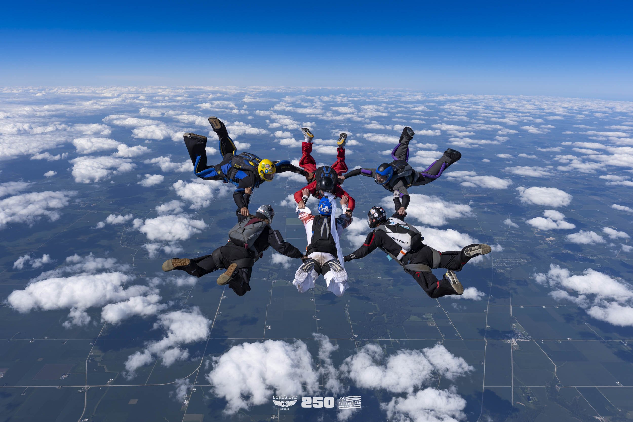 A group of skydivers holding hands in a circle during freefall high above the clouds, with a clear blue sky and a patchwork of farmland below.