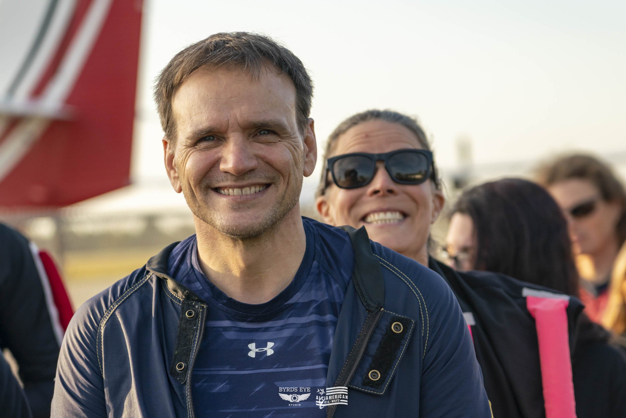 Smiling man in athletic clothing standing outdoors with a group of people, some wearing sunglasses, at what appears to be an outdoor event or gathering.
