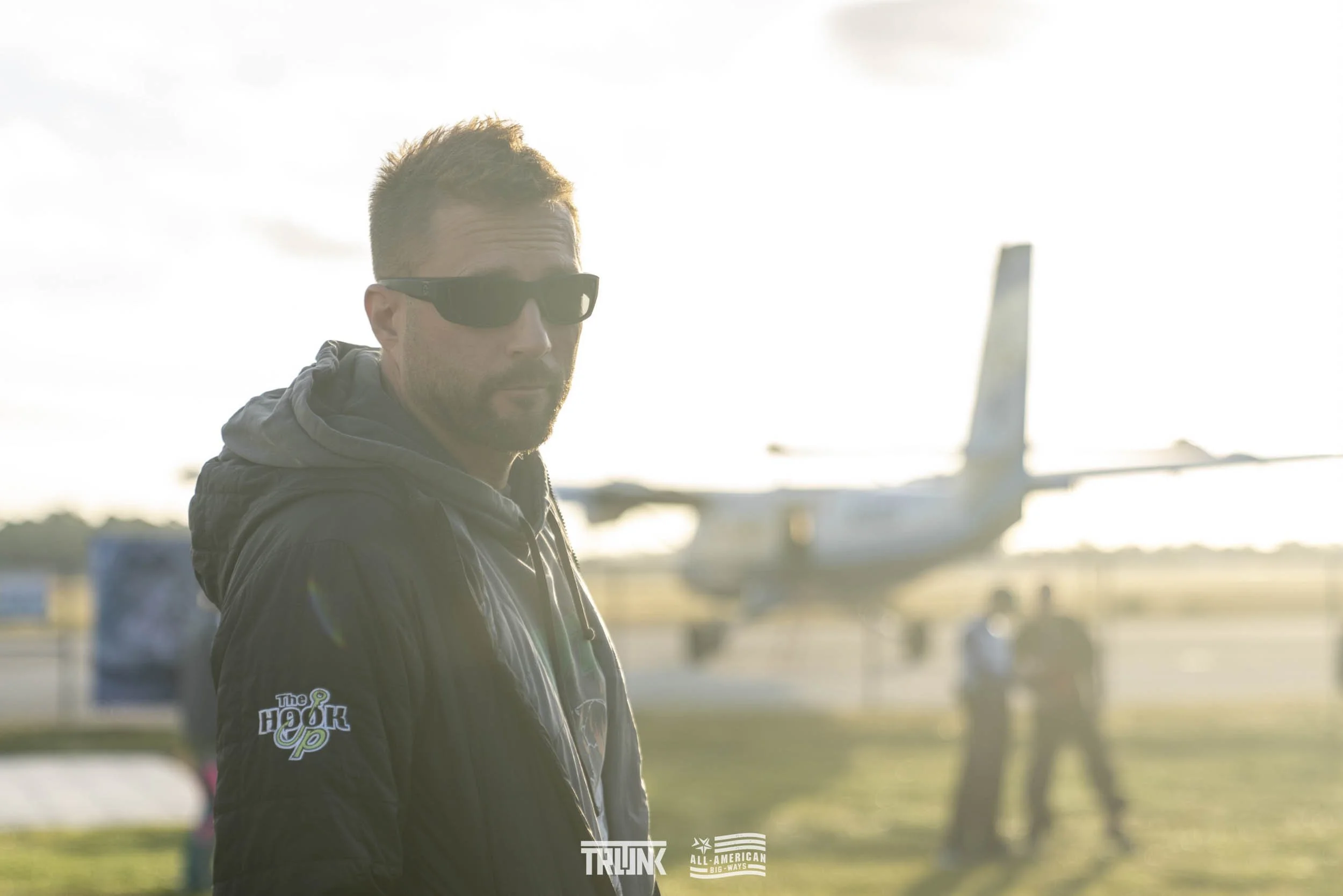 A man with light skin, dark sunglasses, and short brown hair, wearing a black jacket, is standing outdoors with an airplane in the background. The photo appears to be taken during sunset or sunrise with bright backlight.