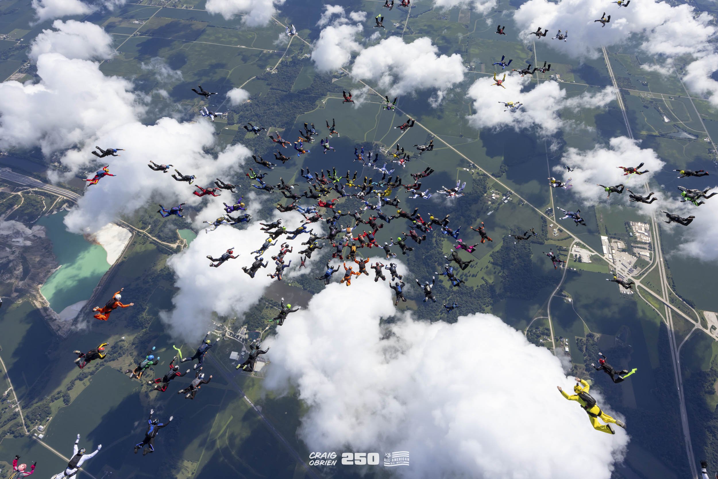 A large group of skydivers in colorful jumpsuits falling through the sky among clouds over a landscape with lakes, roads, and fields.