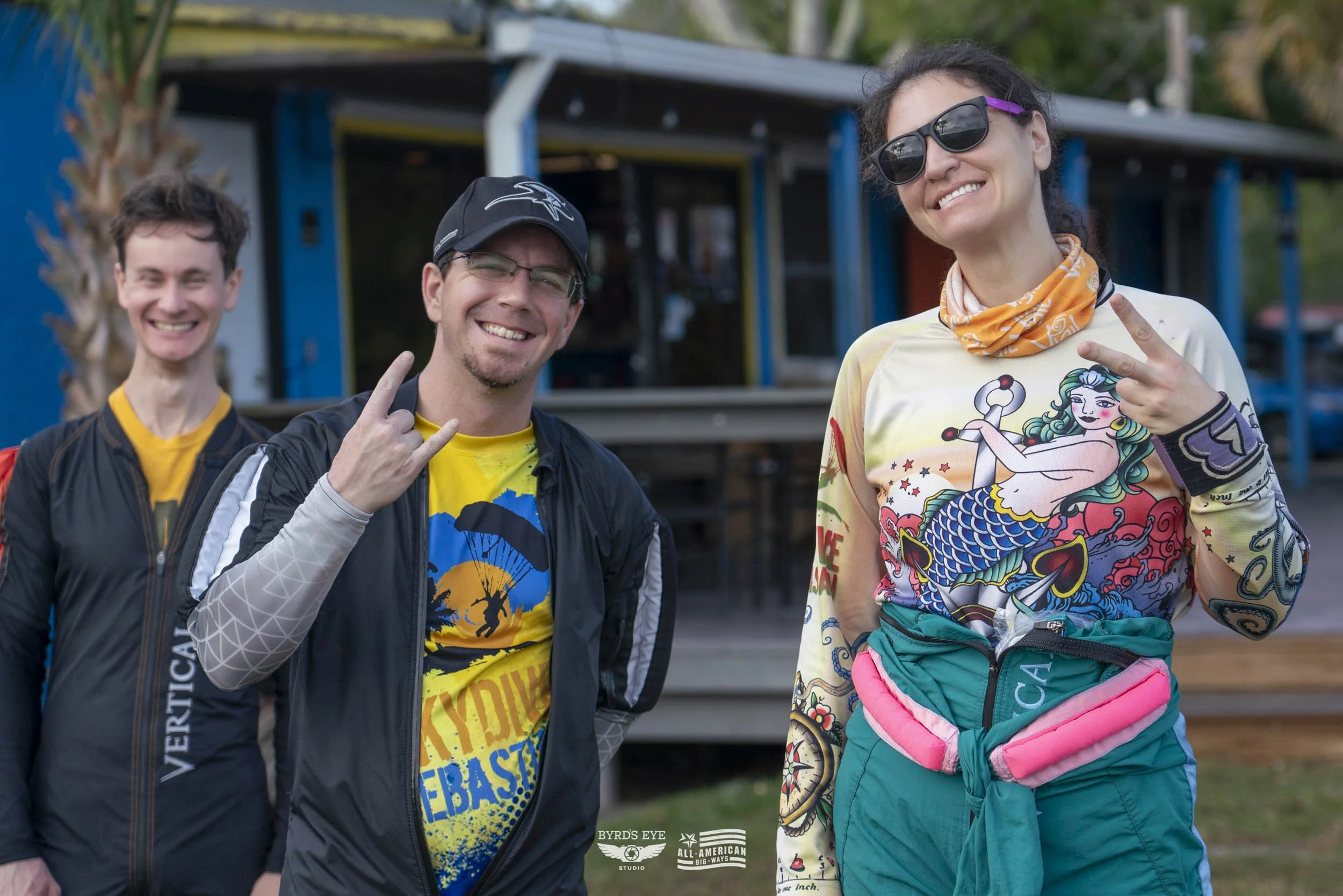 Three friends smiling and making peace signs outdoors, with a colorful building behind them. One woman wears sunglasses and a salmon-colored shirt with a mermaid design; two men wear jackets and T-shirts, one with a kayak or outdoor adventure theme.