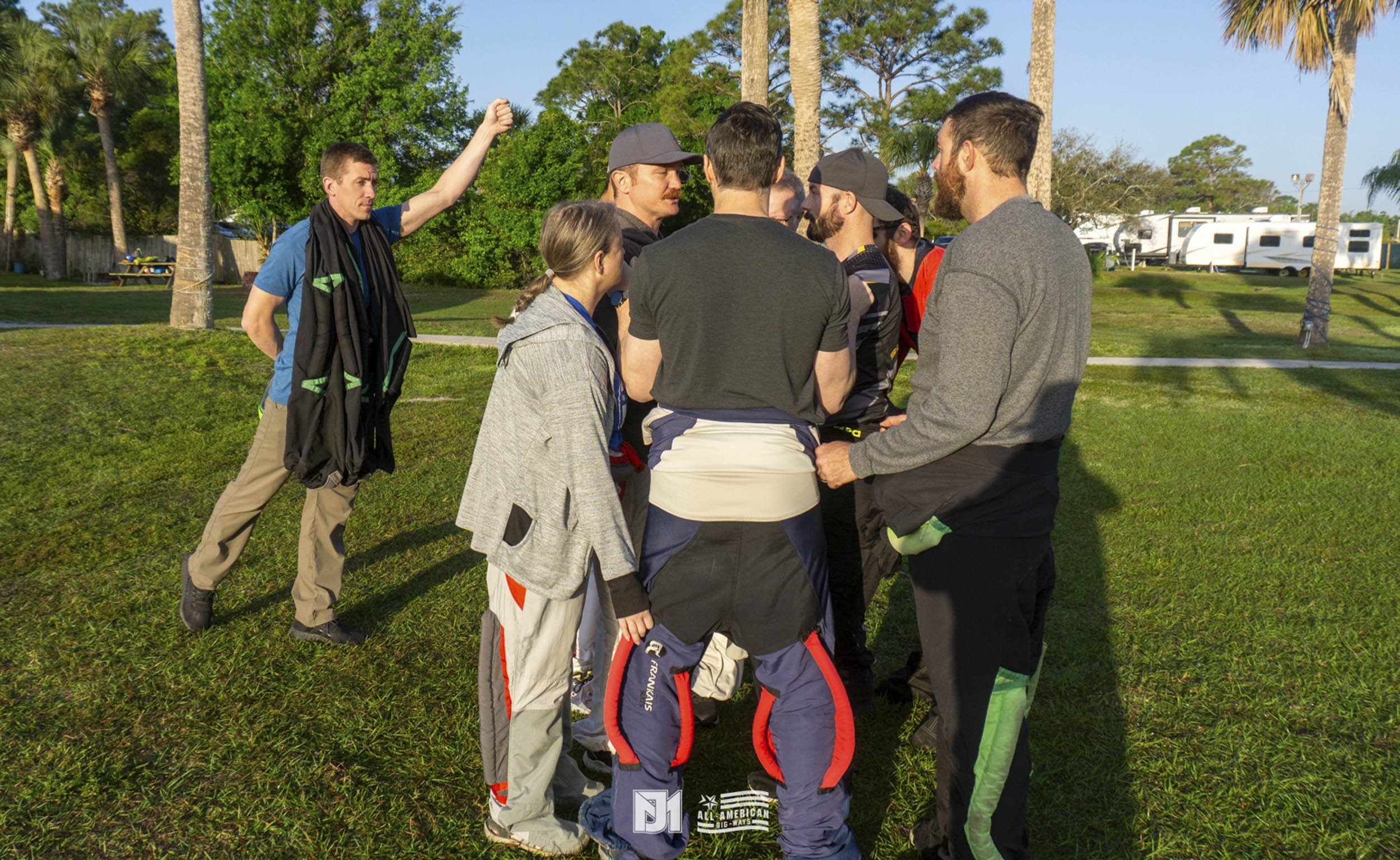 Group of people gathered in a circle on a grassy field outdoors, with some wearing racing gear and others in casual clothes, surrounded by trees and parked trailers or RVs.