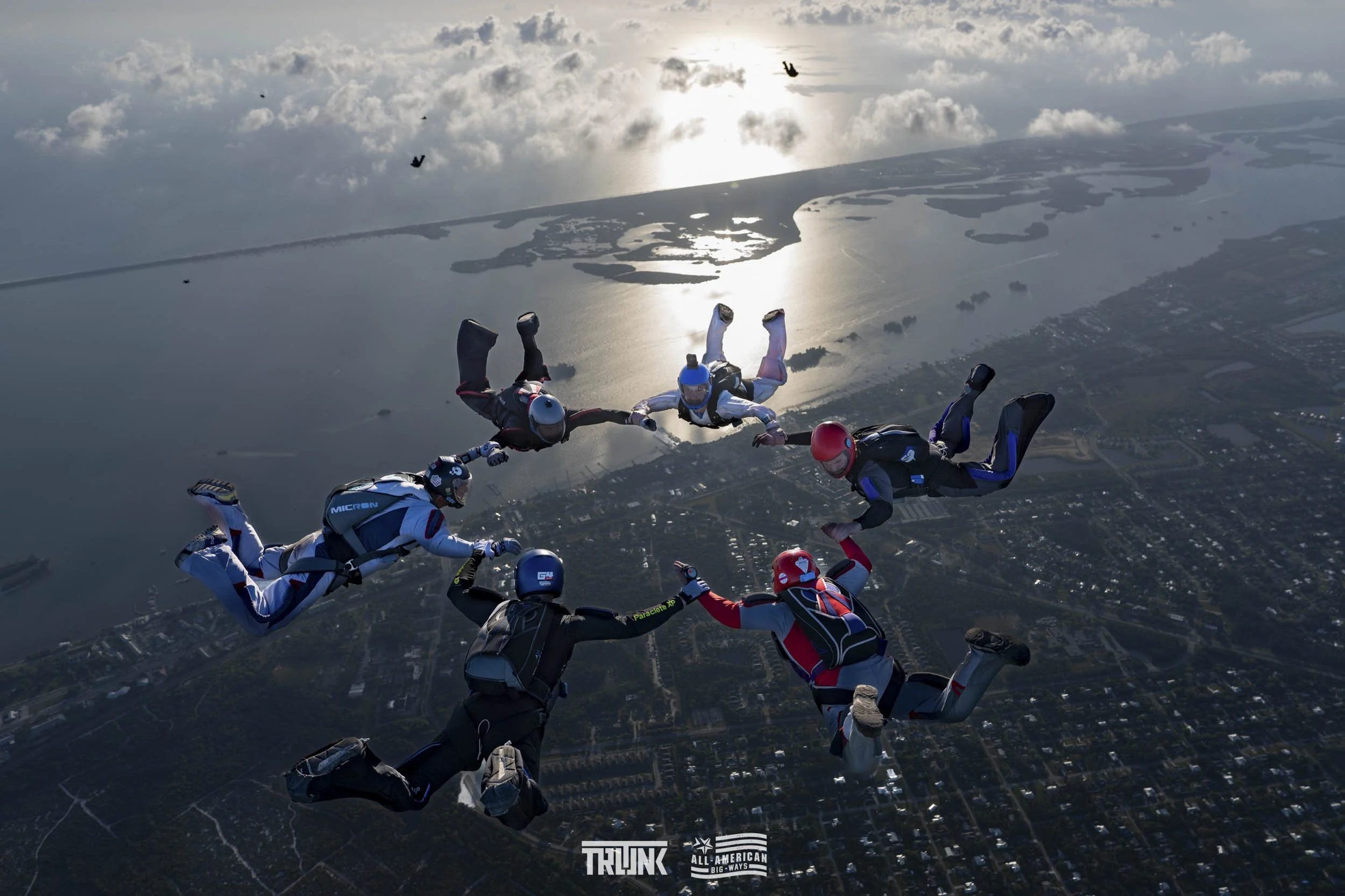 A group of skydivers holding hands in a circle while free-flying above a landscape with water and land, with clouds and the sun reflecting on the water below.