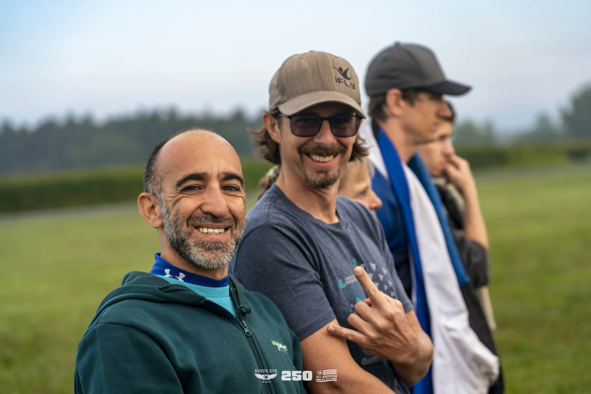 Group of people standing outdoors in a line, with the closest man smiling at the camera. He has a beard and is wearing a dark green hoodie. The man next to him is smiling, wearing a cap and sunglasses, and making a hand sign. Other people in the back
