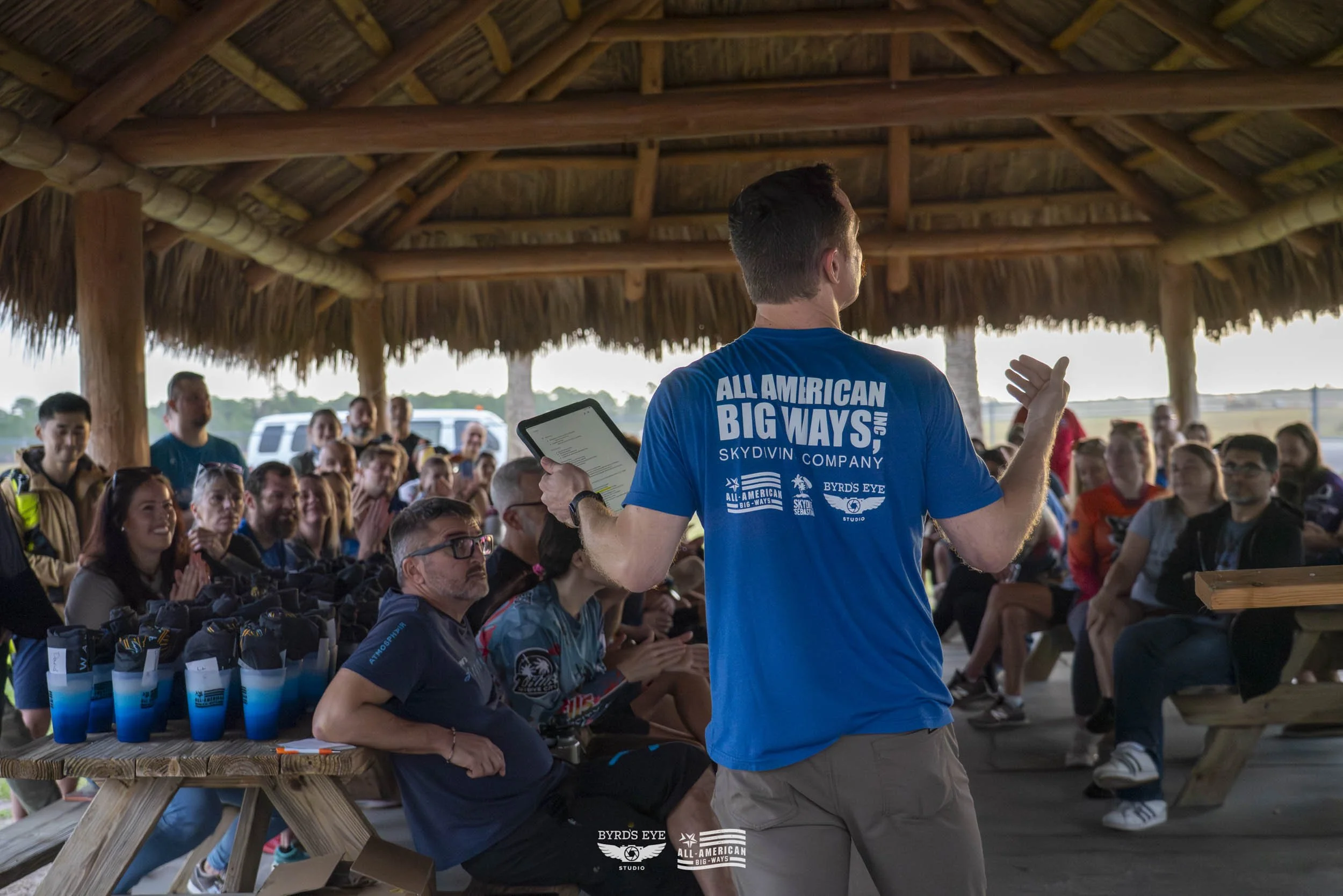 A man in a blue t-shirt giving a presentation to a seated crowd under a thatched open-air structure.