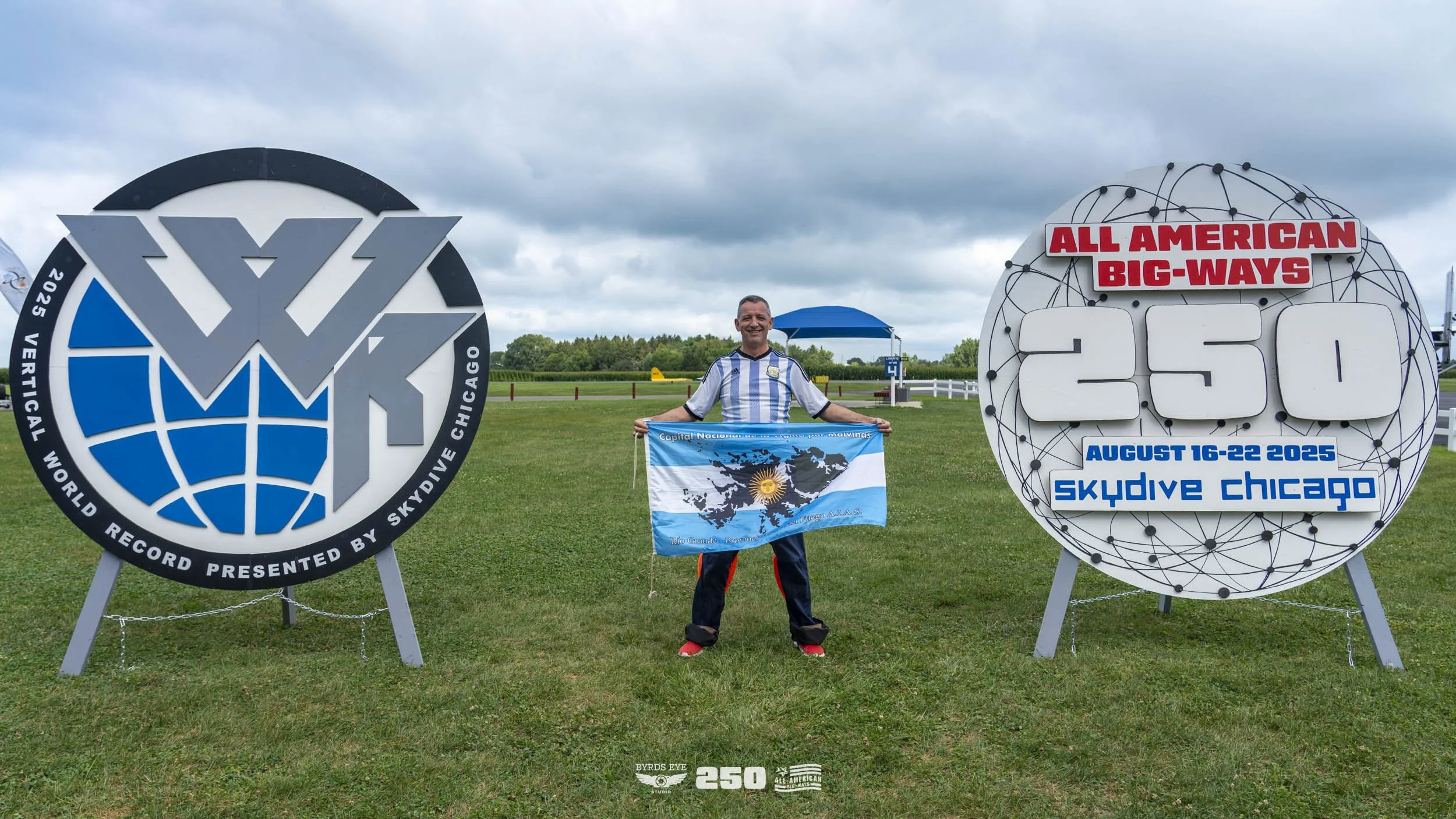 A man holding an Argentine flag stands between two large signs celebrating a world record for high-altitude skydiving. The left sign features a logo of a globe with the words 'World Record Presented by Skydive Chicago.' The right sign states 'All Ame