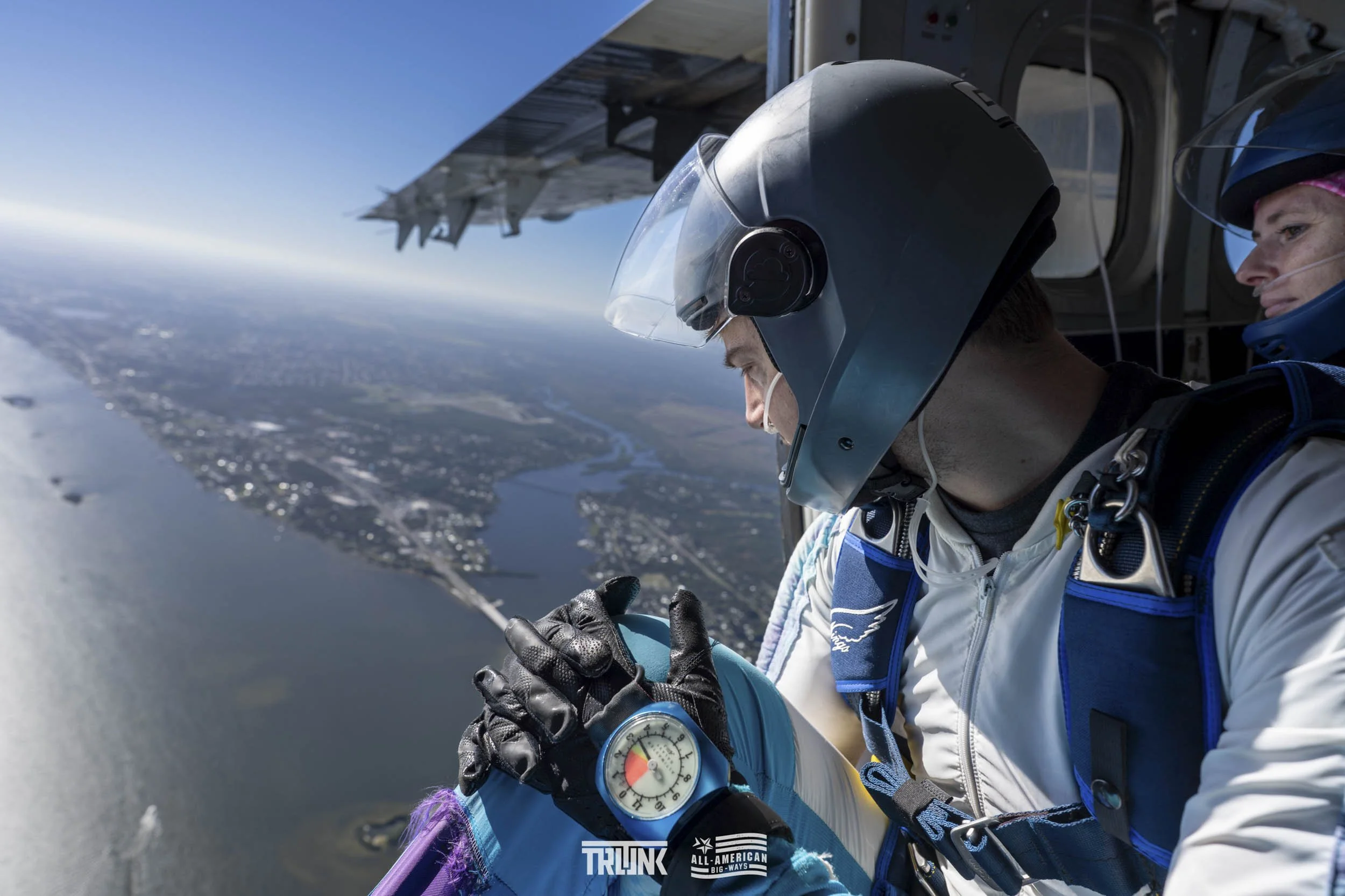 Skydiver looking at an altimeter during a jump, with an ocean and shoreline visible below, and another skydiver in the background.