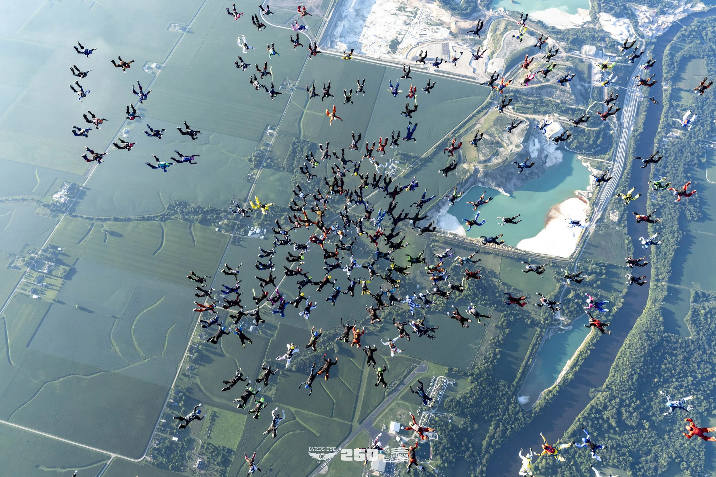 A group of skydivers in colorful jumpsuits free-falling over a landscape of green fields, roads, and water bodies. The skydivers are in a coordinated formation, with some in a circle and others scattered.