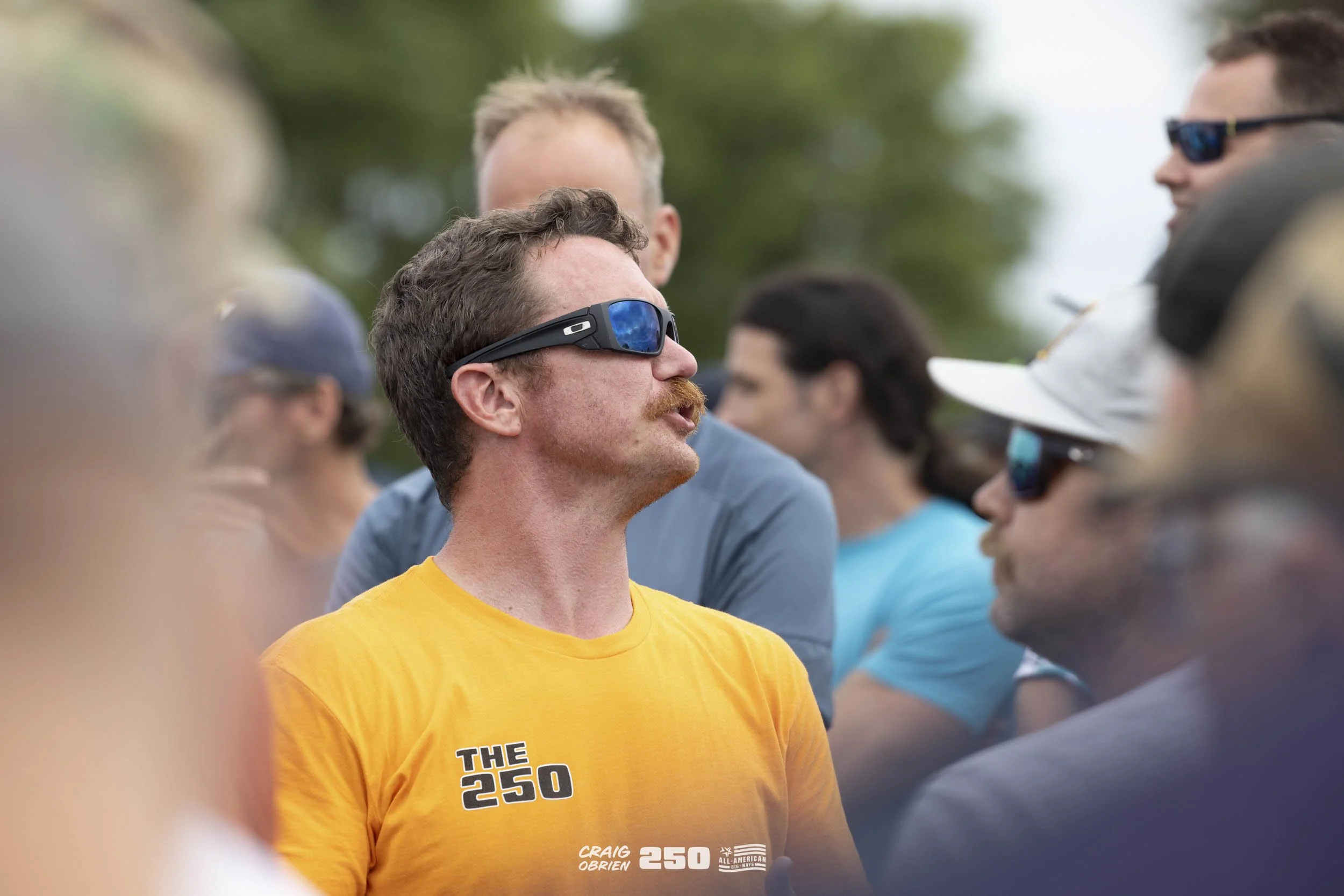 Group of people outdoors, some wearing sunglasses, with a man in a yellow shirt with 'THE 250' and 'CRAIG O'BRIEN' text, standing in profile with a serious expression.