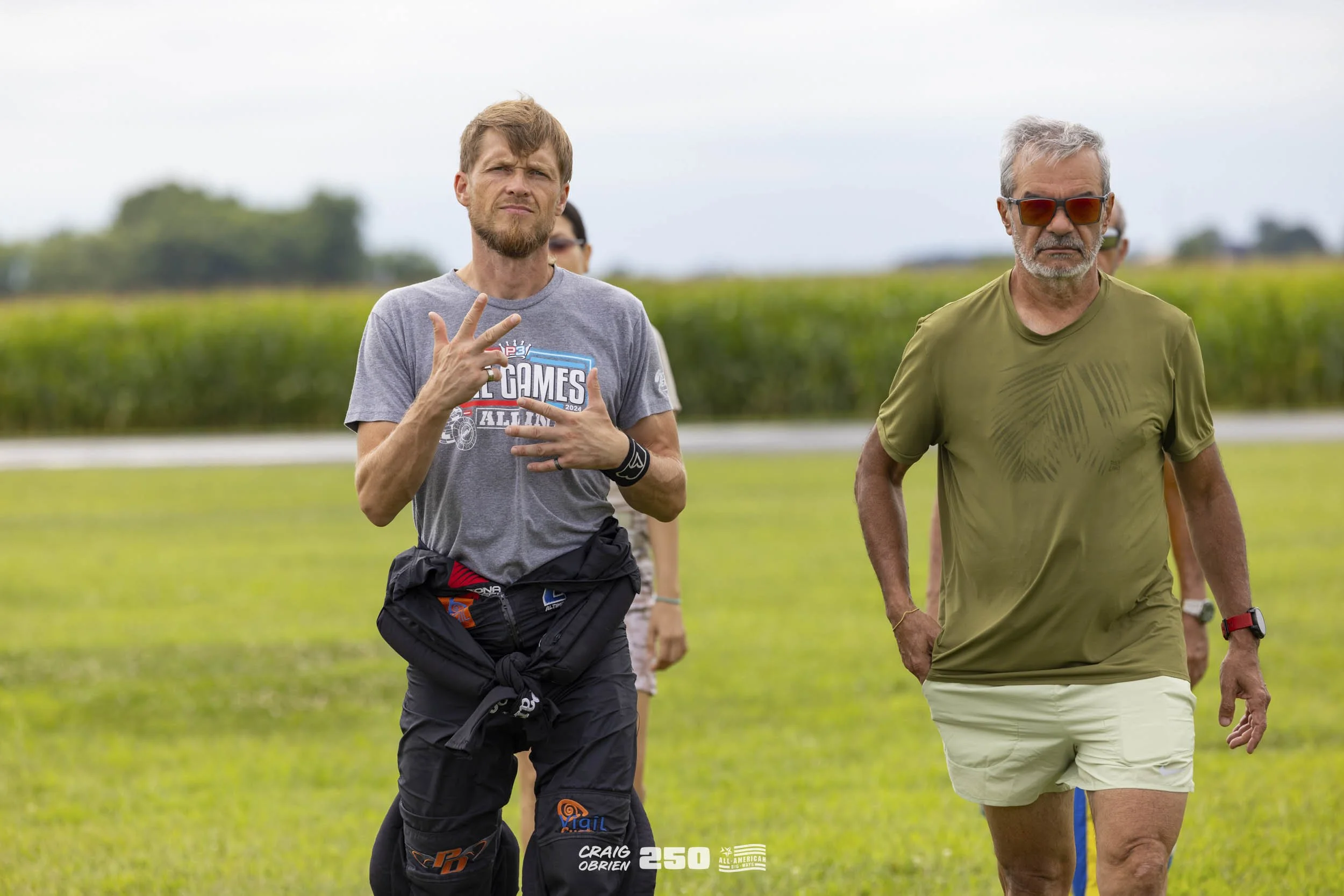 Two men walking outdoors on a grassy field, one wearing a gray T-shirt with a graphic, black pants, and the other in a green shirt and beige shorts, with a blurred background of green landscape.